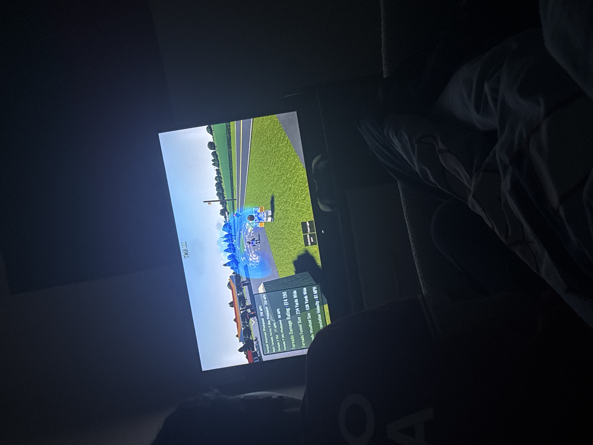 The image shows a television screen displaying a video game, presumably about tornadoes, as indicated by the on-screen text. The in-game environment features green, grassy areas, roads, buildings, and a tornado in progress, represented by blue and white swirling effects. Text on the screen provides detailed statistics about the tornado, including its maximum wind speed, damage rating, funnel size, windfield size, and translation velocity. The time "11:23 AM" is displayed at the top of the screen. The photograph appears to have been taken in a dimly lit room, with part of a person's body visible in the foreground, including a gray top with a partial letter "O". The scene is located indoors, and the information available in the picture does not provide any details about the city of Welshpool in the United Kingdom.