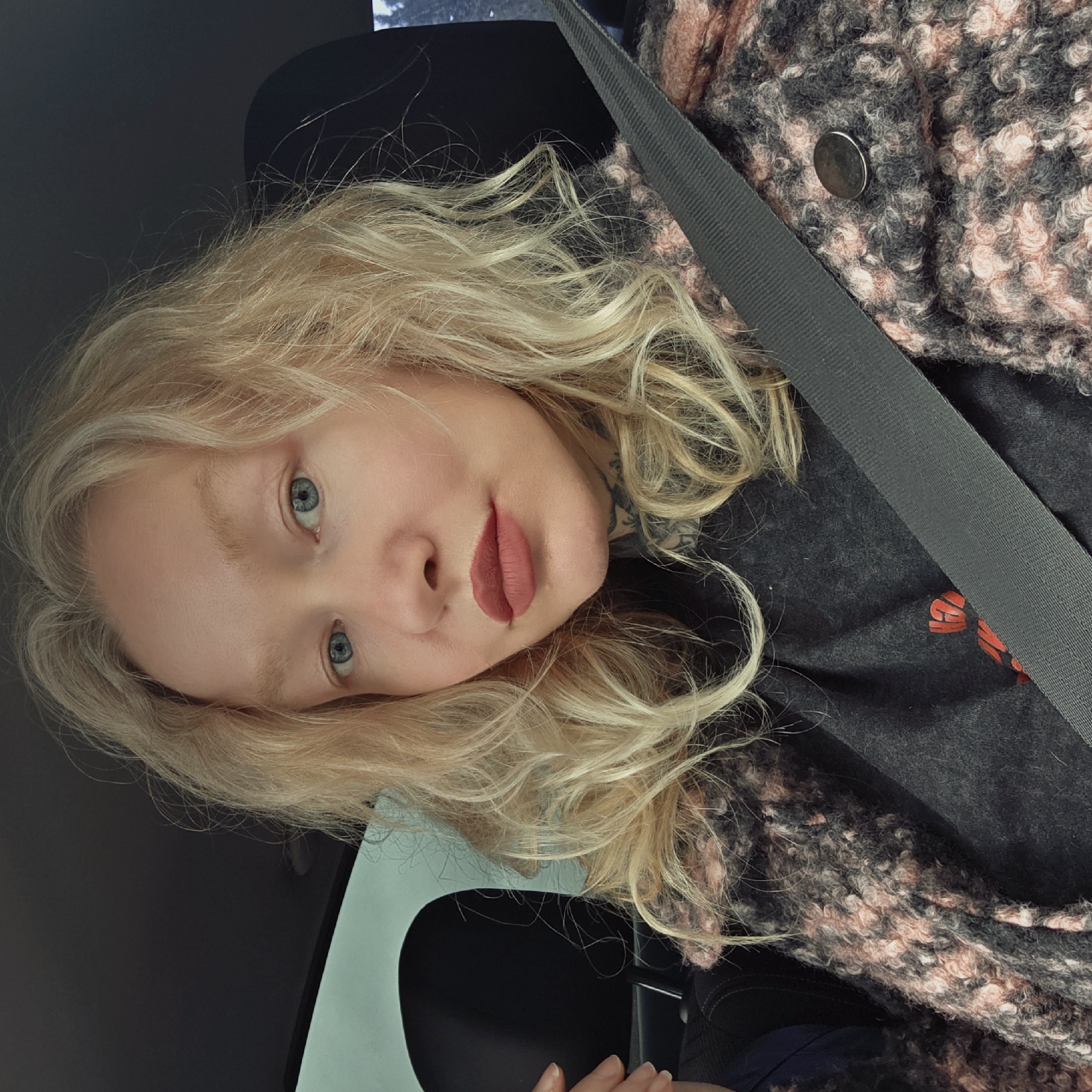 A fair-skinned person with blonde, wavy hair and blue eyes is visible. They are wearing a gray shirt and a multicolored jacket with a button. A gray seatbelt crosses their torso. They are inside a vehicle. The location and city of Gubanitsi, Russia, are not identifiable from the image.