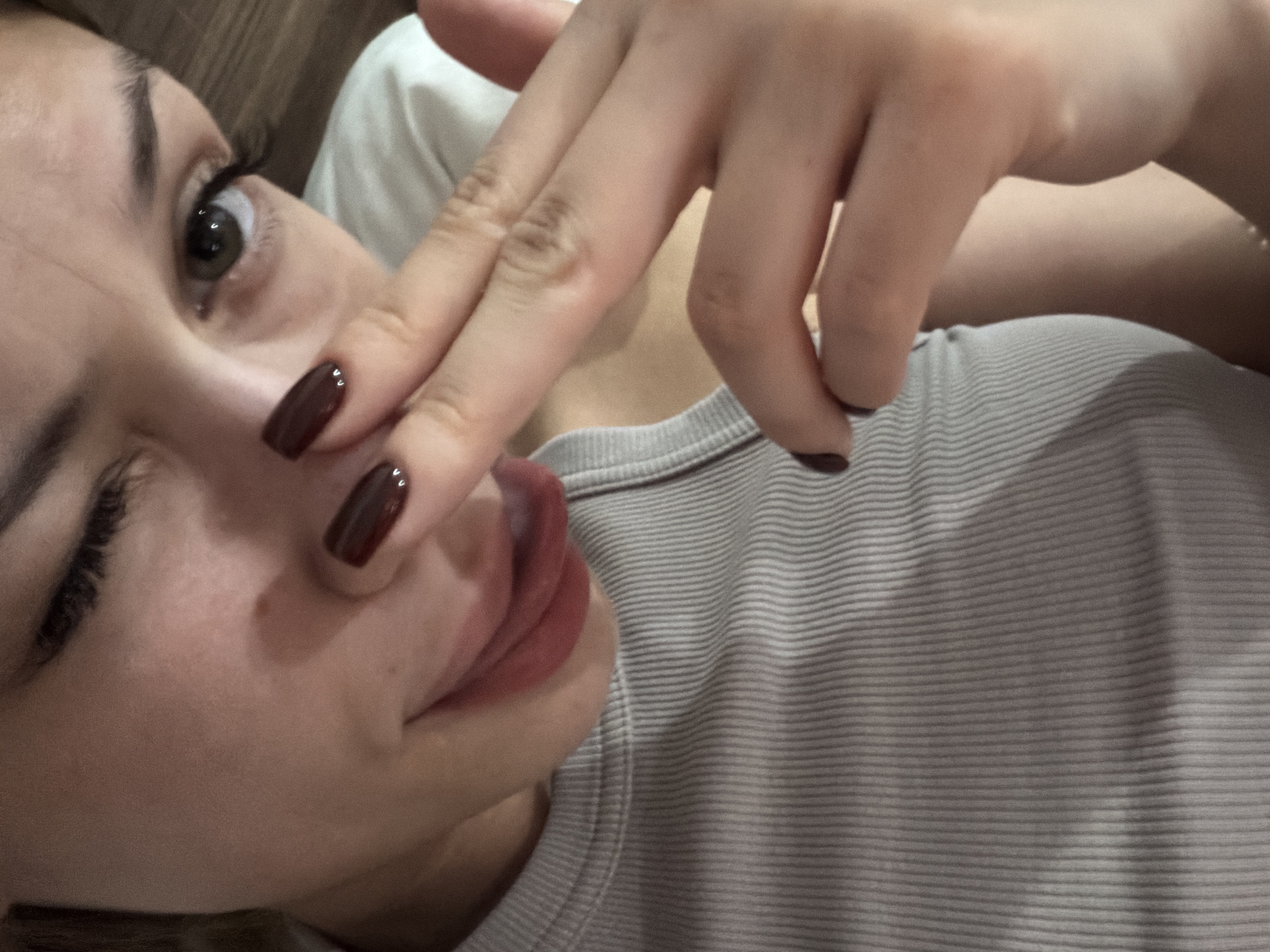 The image is a close-up selfie of a young woman. She is wearing a gray ribbed tank top. Her left eye is open, while her right eye is closed in a wink. Her right hand is raised, with two fingers extended towards her face, touching her nose. Her lips are slightly parted, with her tongue visible. She has dark, polished fingernails. The background appears to be a wooden surface, possibly furniture or a wall. There is no discernible location in the city of Đà Nẵng, Vietnam.