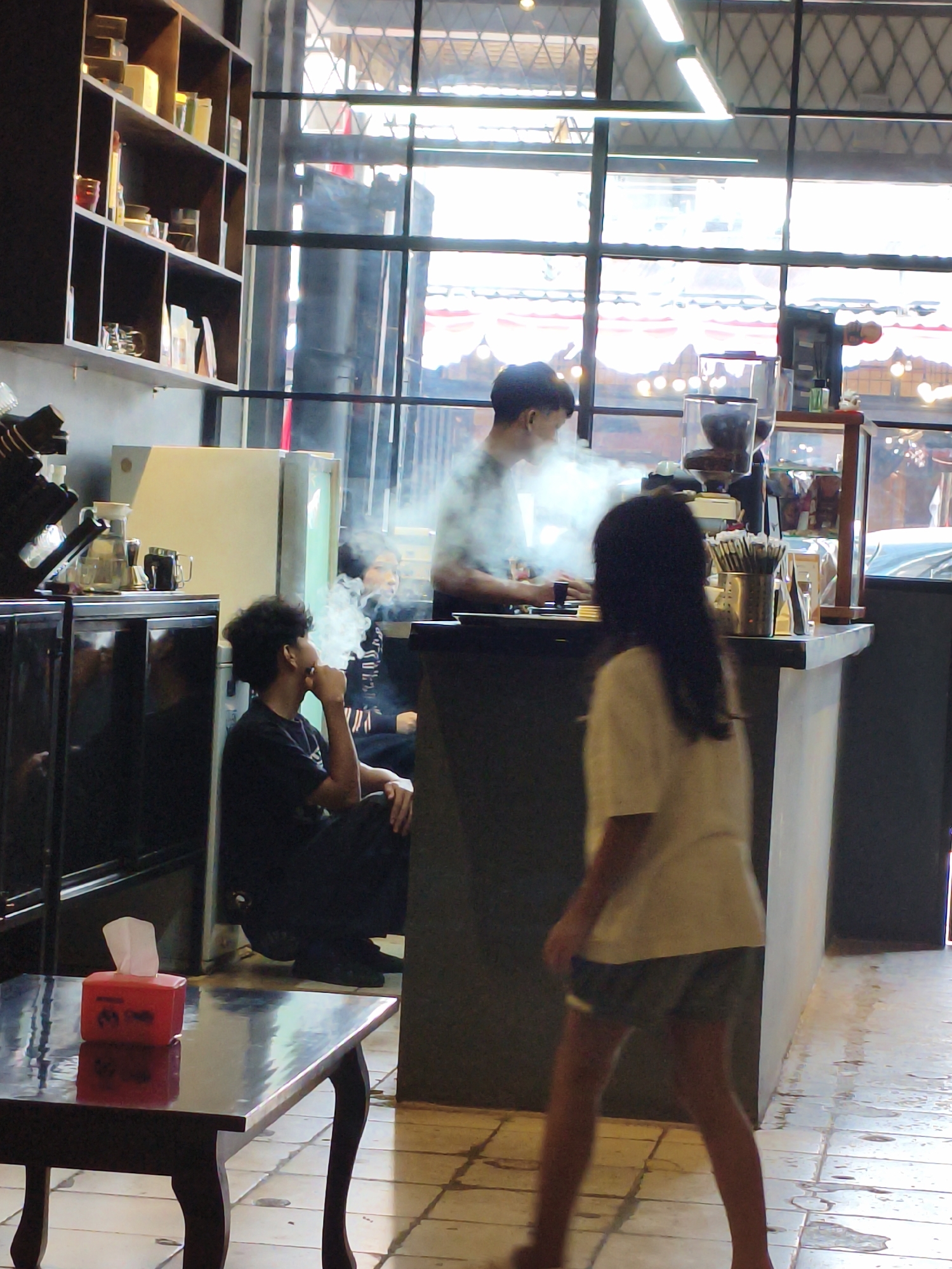 The image shows the interior of a cafe, likely in the City of Tebing Tinggi, Indonesia, during daylight hours. A man is sitting on the floor and vaping, with a cloud of vapor rising around him. A woman with long dark hair is walking away from the camera, appearing to be a customer. Behind the counter, another person is visible, possibly a barista, and there are coffee making machines and shelves stocked with items. In the foreground, a dark wooden table with a red tissue box is visible. The cafe has large windows looking out onto a street scene, and the interior features tiled flooring and fluorescent lighting.