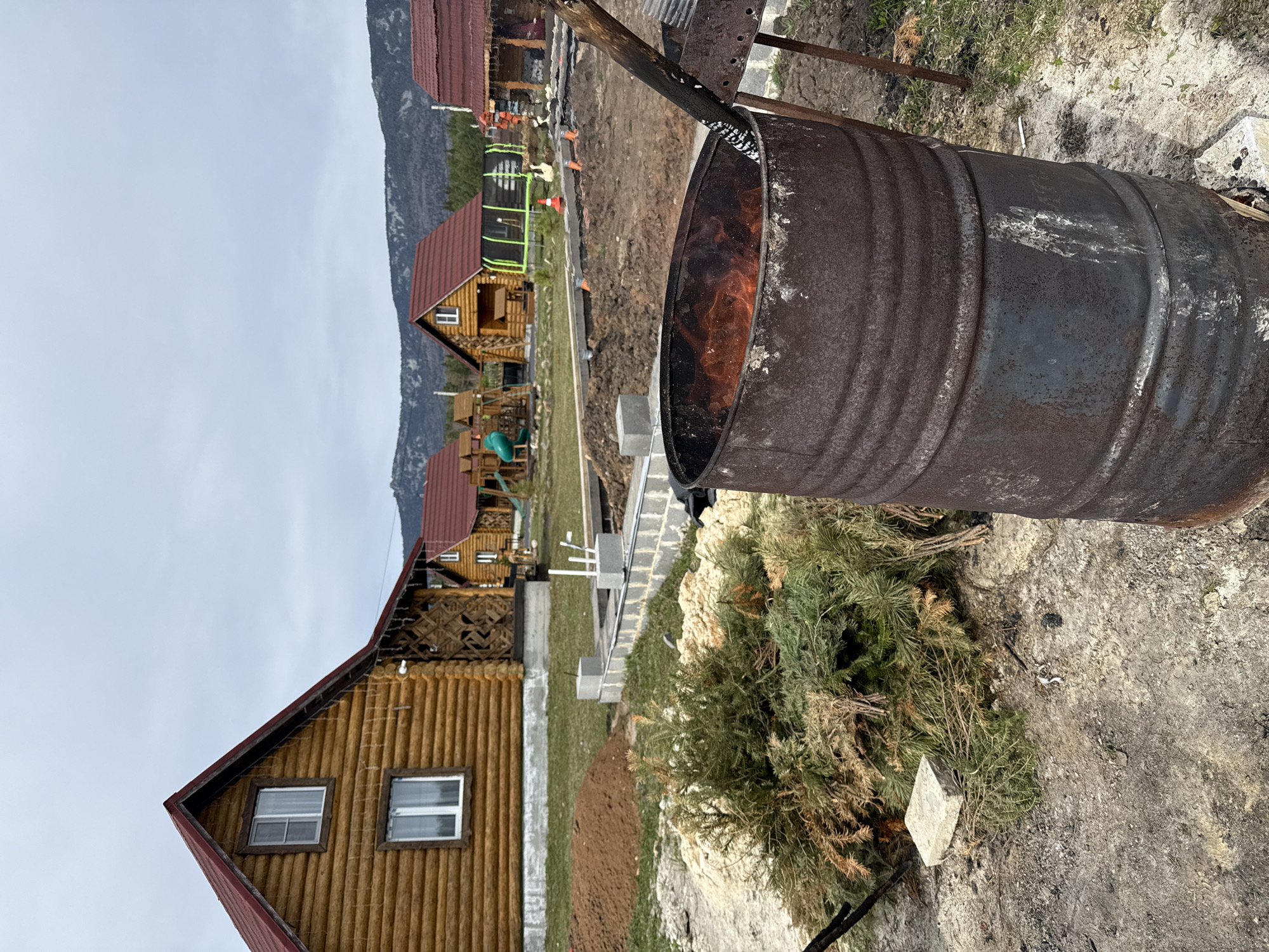 The image depicts a rustic outdoor scene in the Tyulyuk region of Russia. In the foreground, a heavily rusted and burnt metal barrel sits, its interior filled with flames. The ground around the barrel is a mix of bare earth, dried grass, and scattered pine needles. A discarded Christmas tree lies near the barrel, and a small concrete block is partially obscured by the tree’s branches.
In the background, a row of log cabins with red roofs stretches into the distance. These cabins appear to be part of a resort or vacation complex. Between the barrel and the cabins, there is a construction site with freshly laid concrete blocks and other building materials. A trampoline and other recreational equipment are visible among the cabins, suggesting a leisure-oriented environment.
The sky is overcast, and a mountain range is visible in the far distance. No people are visible in the image. The scene suggests a cold climate and a rural or semi-rural setting. The burning barrel and the construction site imply ongoing activity and potential development in the area.