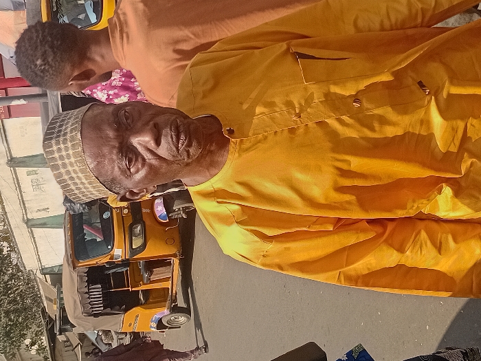 An older man wearing a patterned cap and yellow shirt stands in front of yellow auto rickshaws. Another person is partially visible behind him, wearing an orange shirt with a floral-patterned section. The setting appears to be outdoors, possibly a street or market area. The sunlight is bright, creating shadows on the subjects and surrounding area.