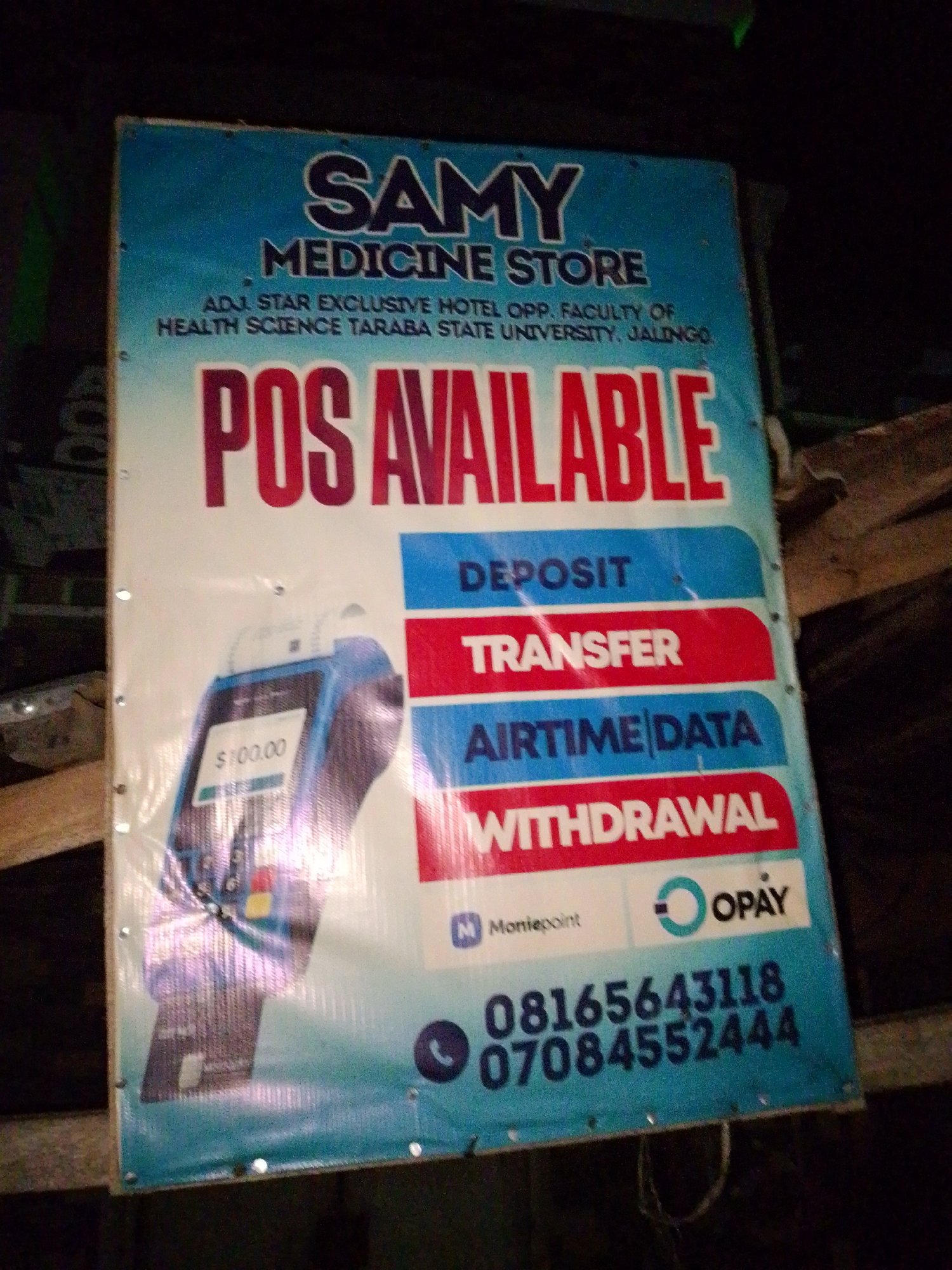 The image shows a banner for "SAMY MEDICINE STORE" located in Jalingo, Nigeria. The banner advertises "POS AVAILABLE" for deposit, transfer, airtime, data, and withdrawal services. The store's address is listed as "ADJ. STAR EXCLUSIVE HOTEL OPP. FACULTY OF HEALTH SCIENCE TARABA STATE UNIVERSITY, JALINGO." Below this, there are logos for Moniepoint and OPay, and phone numbers: 08165643118 and 07084552444. A POS machine is depicted on the left side of the banner. The banner is hung outdoors, possibly on a wooden structure, and the lighting suggests it is nighttime.