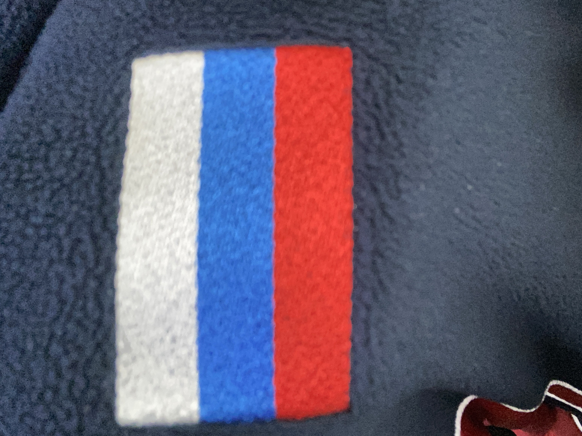 The image shows a close-up of a stitched patch depicting the Russian flag. The flag has three horizontal stripes: white on top, blue in the middle, and red on the bottom. The flag is attached to a dark blue fabric, possibly part of a garment or textile. There are no people or any environmental context visible that would indicate a specific location in Luzhitsy, Russia. The focus is solely on the flag patch.