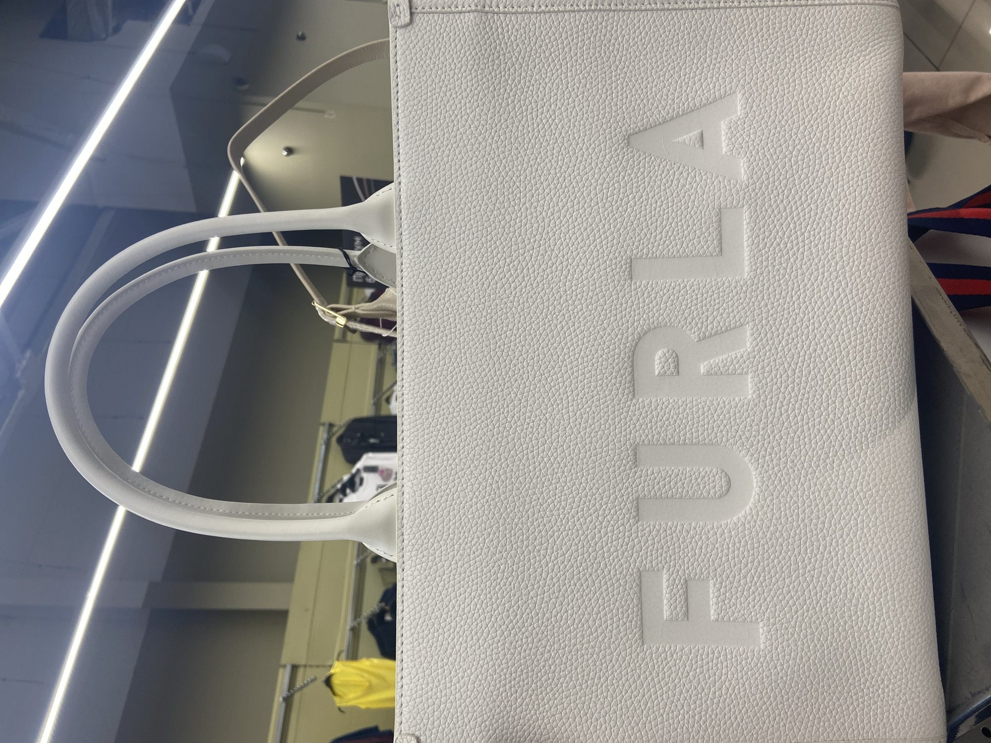 The image shows a white leather tote bag with the embossed word "FURIA" on it. The bag has two top handles and a detachable shoulder strap. It is displayed in a retail environment, with clothing racks visible in the background. The lighting suggests an indoor setting, likely a store. There is no visible indication of the specific city or location, including Magnitogorsk, Russia. No people are clearly visible in the foreground.
