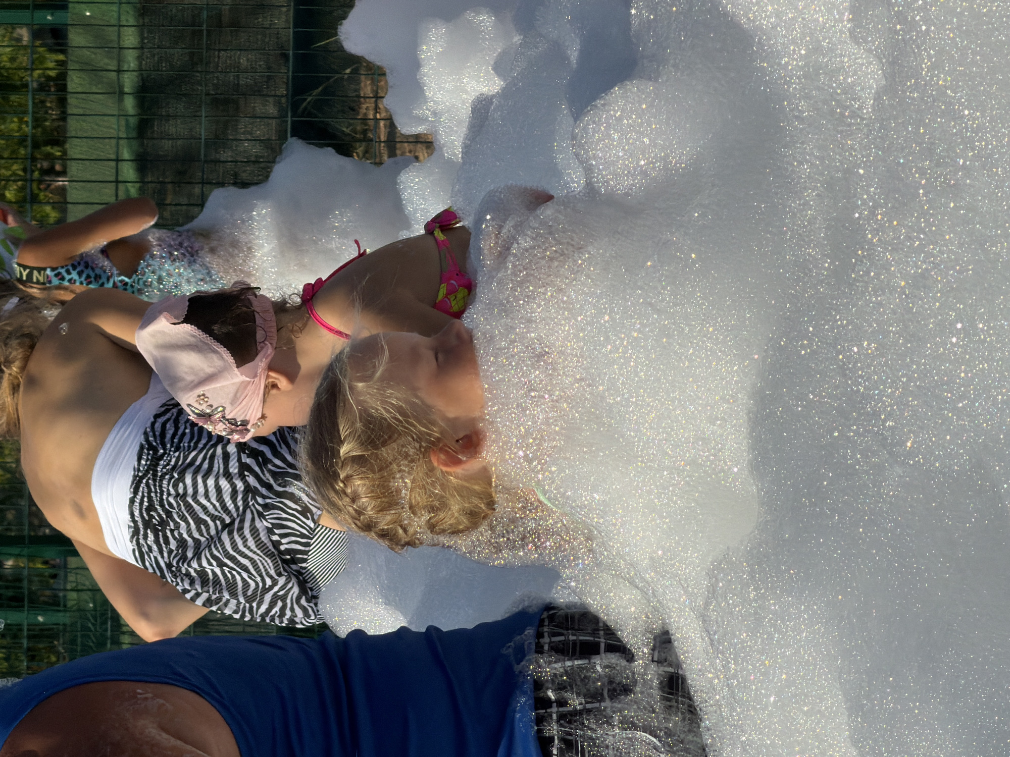 The image shows two young girls playing in a large pile of white foam, likely from a foam party. The girl on the left is wearing a white swimsuit and a pink headband with floral embellishments. She is lying on her back and her face is partially obscured. The girl on the right is wearing a pink and yellow bikini. Her head is tilted back, and her blonde hair is styled in a braid. Both girls appear to be enjoying themselves in the bubbly environment.
The scene is outdoors, with a green metal fence visible in the background. Sunlight is reflecting off the foam, creating a sparkling effect. The time of day appears to be sunny and warm, suggesting it's likely daytime during pleasant weather. While no specific emotions are explicitly depicted, the context of a foam party implies a fun and playful atmosphere.
There is text visible on the swimsuit of the girl on the left, which appears to say "Y N A". The overall setting suggests a recreational activity, possibly at a park, backyard, or event venue. The foam is so abundant that it covers most of the subjects, giving a sense of immersion and playfulness. The focus of the image is on the children's interaction with the foam.