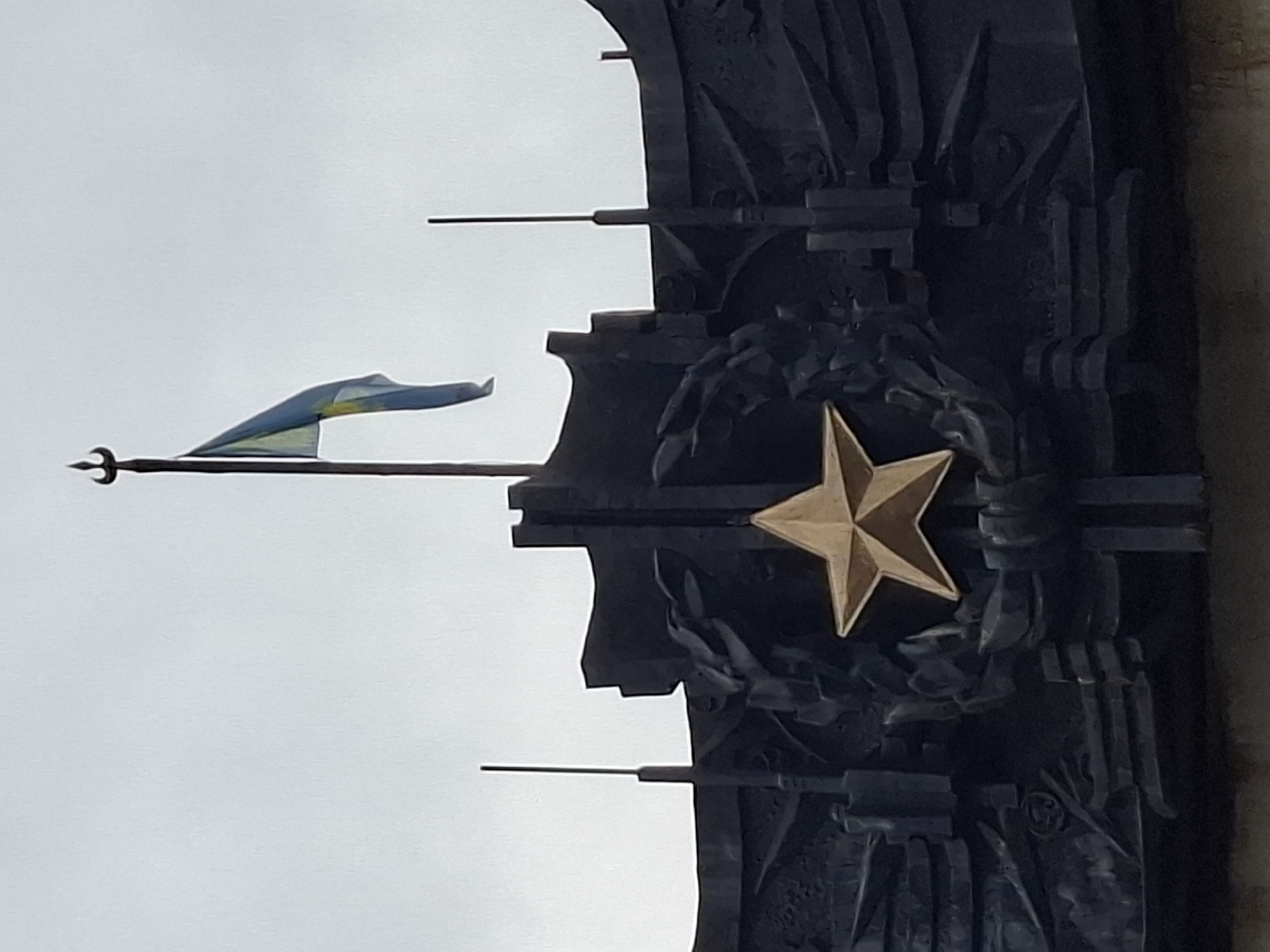 The image shows a close-up view of a monument in Almaty, Kazakhstan. Dominating the center is a large, golden five-pointed star, set within a dark, intricately carved wreath. Above the star, a flag with blue and yellow stripes flies from a flagpole topped with a trident-like ornament. The monument itself is made of dark metal, with abstract, geometric elements and what appear to be stylized representations of figures or objects integrated into its design. The background is a plain, overcast sky. The monument is likely situated outdoors, given the flag and the atmospheric conditions depicted.