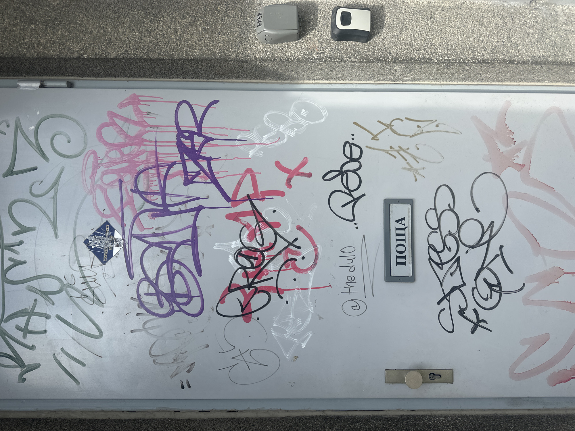 A metal door or hatch is covered with multiple layers of graffiti in various colors, including green, pink, purple, red, black, white, and brown. The door is set into a grey concrete or stone wall. There is a combination lockbox and a key lockbox mounted above the door. A small, rectangular sign with the word "ПОЩА" (which means "Mail" in Bulgarian) is fixed to the door near its center. A door handle is located near the bottom of the door. The location is likely in Varna, Bulgaria, based on the Bulgarian word on the sign.