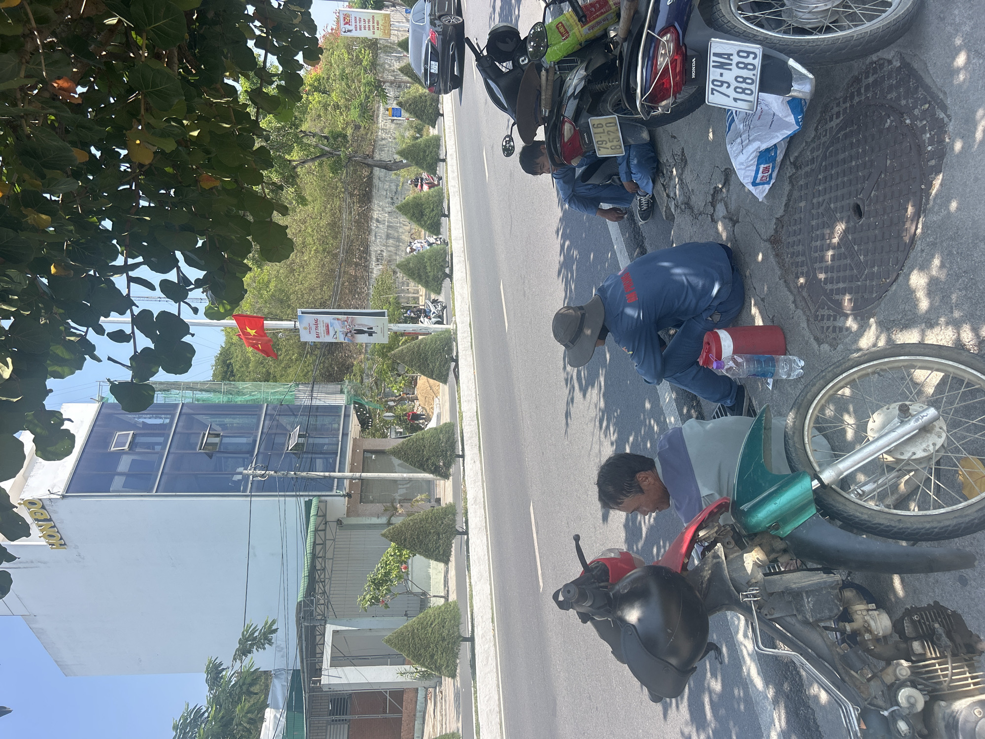 Three men are fixing motorcycles on the side of a road in Bắc Nha Trang, Vietnam. One man in a blue uniform and hat is kneeling near a red and white container and a water bottle, working on a motorcycle. Another man in a blue uniform is kneeling near a motorcycle with a license plate that reads "79-L6 8520." The third man is kneeling near a motorcycle with a red gas tank and a black helmet. A motorcycle nearby has a license plate that reads "79-NA 188.89." The "HONDO" building and a Vietnamese flag on a pole are visible in the background, along with neatly trimmed cone-shaped bushes. Other motorcycles and a car are parked further down the road. The road is cracked and shaded by trees. A manhole cover is on the side of the road.