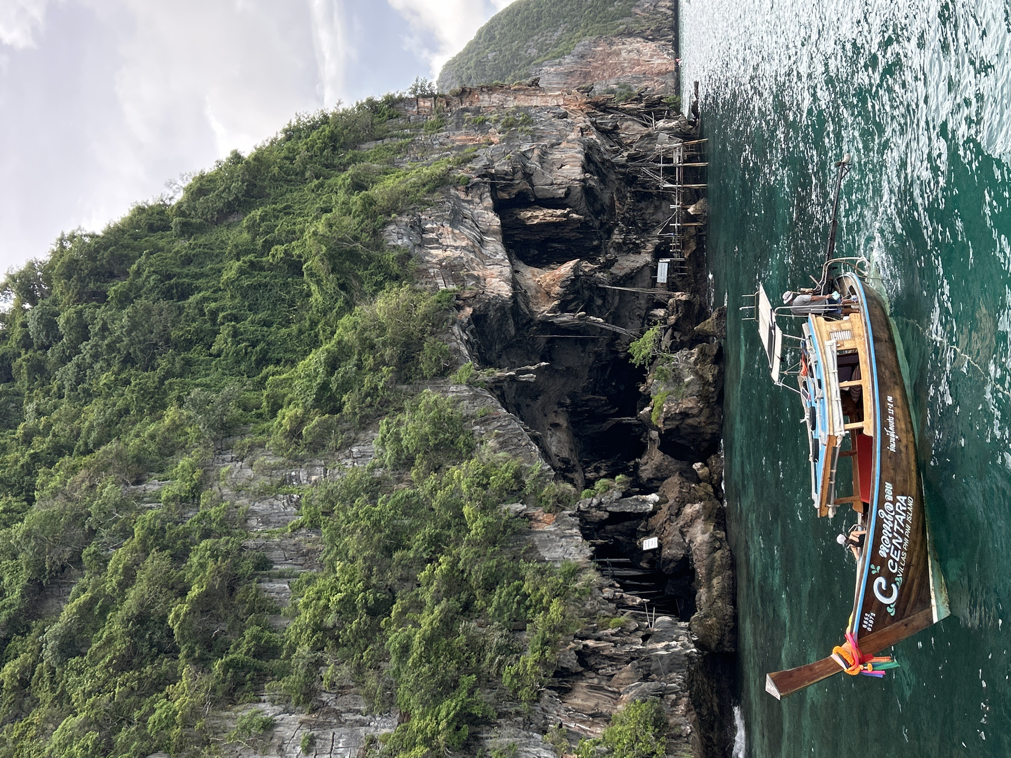 A longtail boat is docked in the turquoise waters of Ao Nang, Thailand, near a limestone cliff face covered in lush green vegetation. The boat, with the name "CENTARA VILLAS PHI PHI ISLAND" emblazoned on its side, has a wooden hull and a covered seating area. A person is visible in the boat, steering it. The cliff features a large cavernous opening and some wooden scaffolding or structures extending from it. The water is clear and appears to be slightly choppy. The sky is overcast with scattered clouds.