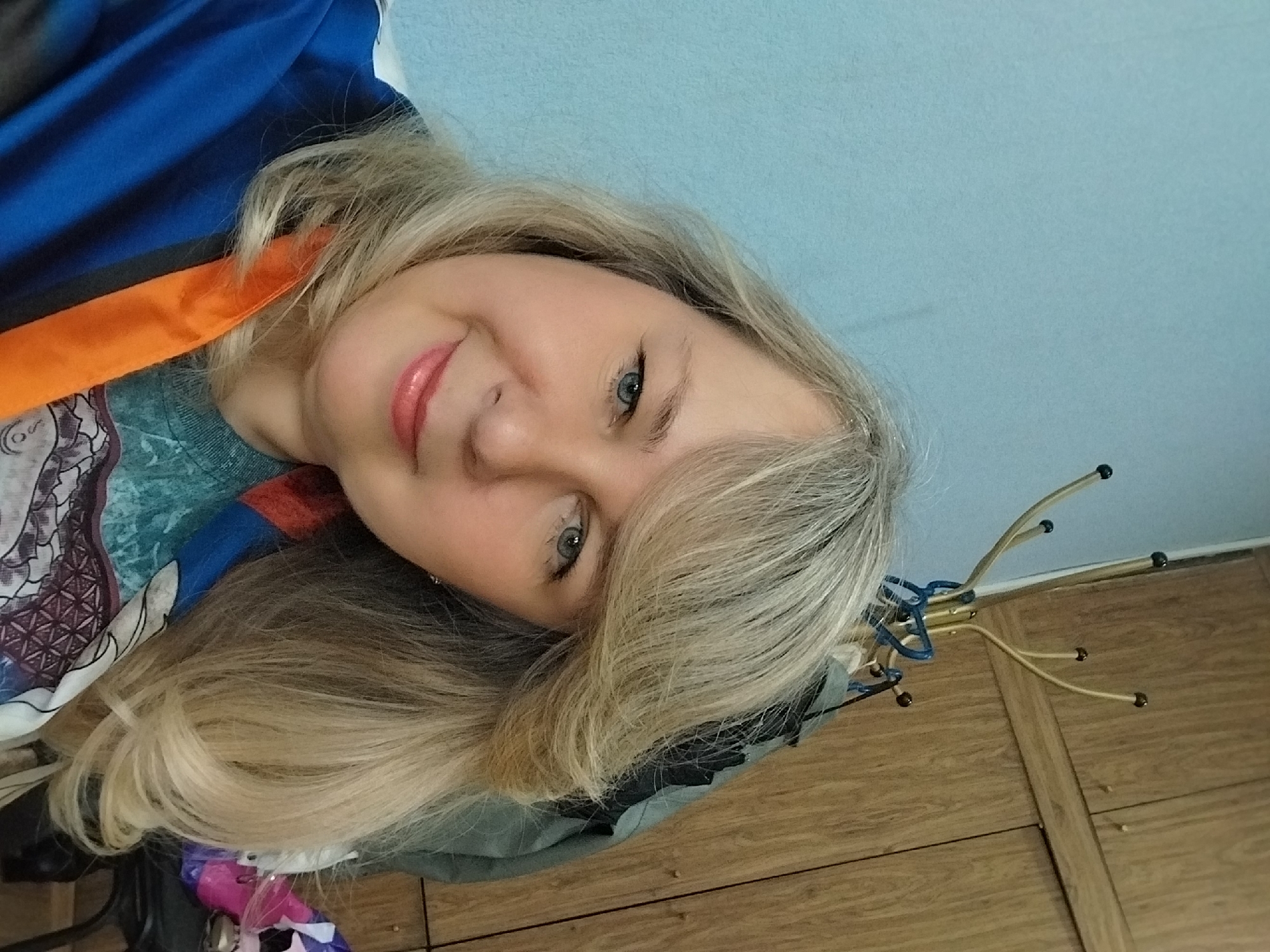 A woman with blonde, shoulder-length hair is posing for a selfie. She is smiling slightly and has blue eyes. She is wearing lipstick and is positioned against a light blue wall. She also has clothes hanging near her and some of it is visible in the photograph. Some of it is blue, orange, gray and purple.
The location is impossible to determine from the visual information provided.