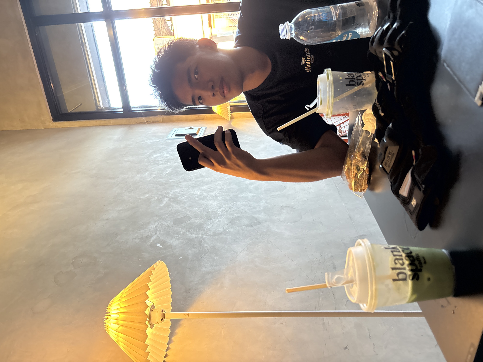 In a dimly lit cafe in Davao City, Philippines, a young man sits at a dark table, illuminated by a yellow pleated lamp on his left and natural light streaming through a large window on his right. He is holding a black smartphone in his right hand, his index finger extended. He wears a black t-shirt with white lettering that reads "Just Motion OXON". In front of him are two disposable cups with straws, one containing a green beverage and the other appearing empty or containing ice. A clear plastic water bottle is to his right. A sandwich wrapped in plastic and some black fabric items, possibly gloves, are also on the table. The wall behind him is textured and grey, with the lamp casting a warm glow. The overall atmosphere is casual and relaxed.