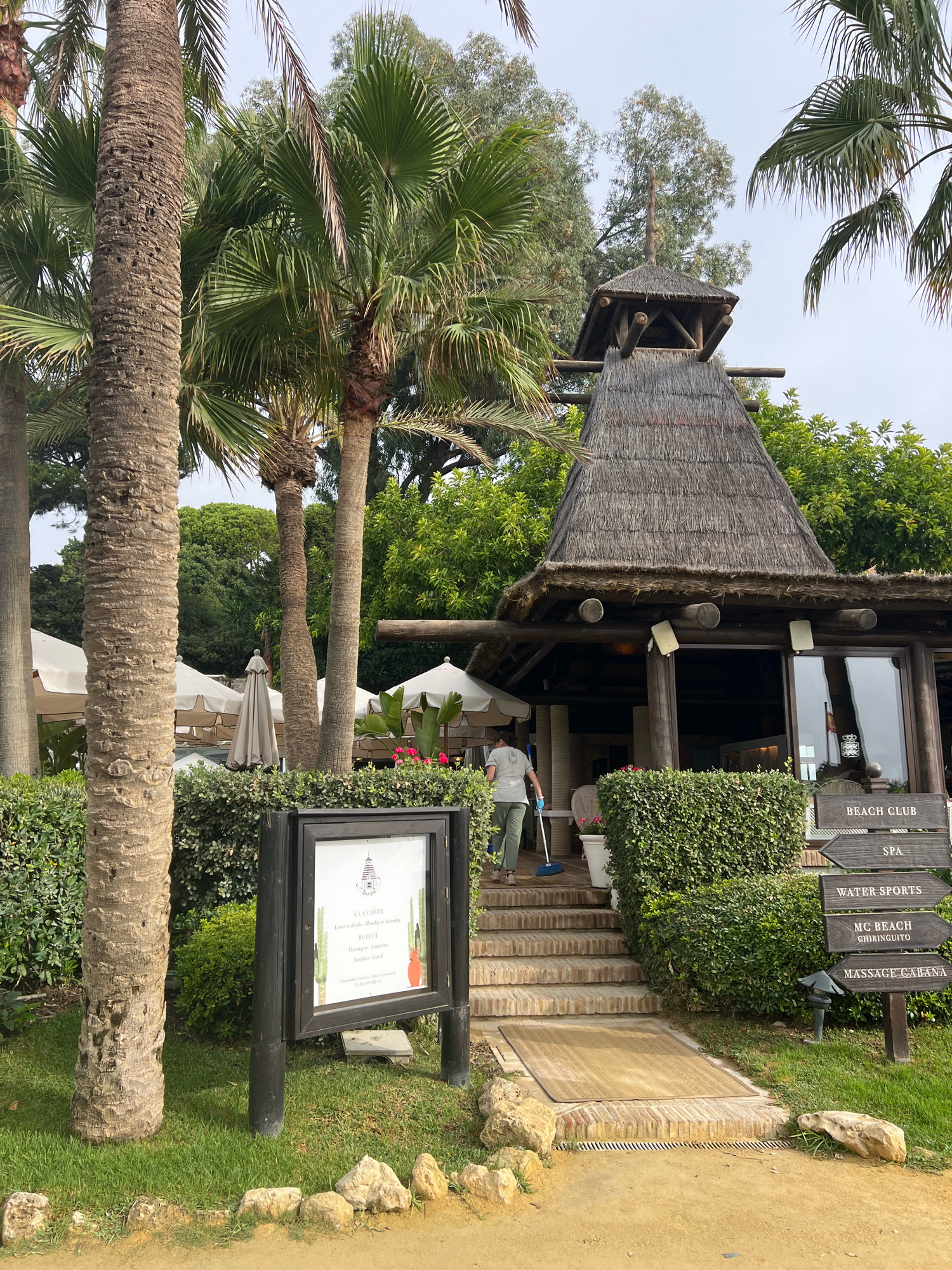 The image depicts a tropical outdoor scene, likely a resort or beach club, with prominent palm trees and lush greenery. A person is sweeping the steps leading up to a building with a thatched roof. The building appears to be part of a beach club, indicated by signage indicating "BEACH CLUB," "SPA," "WATER SPORTS," "MC BEACH GHIRINGUITO," and "MASSAGE CABANA." The setting suggests a warm climate, consistent with Marbella, Spain, known for its coastal resorts. The overall atmosphere is one of a relaxing, vacation-oriented environment.