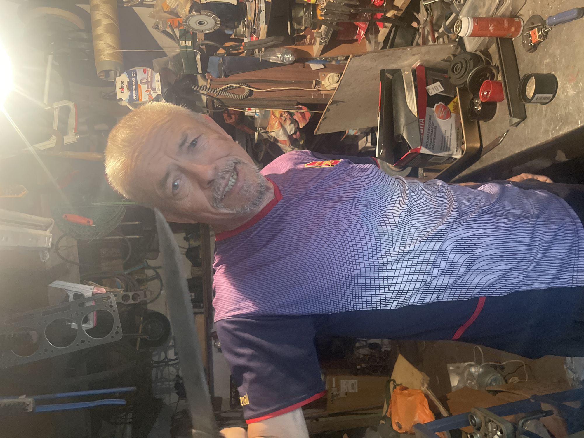 An older Caucasian man with short, blonde hair and a mustache smiles at the camera in a cluttered workshop. He is wearing a purple and blue t-shirt with a geometric pattern and red trim around the neck and sleeves. He is holding a long, flat object, possibly a tool or a piece of metal, in his left hand, angled towards the viewer. The workshop is filled with various tools, equipment, and supplies, including shelves stacked with items, electrical cords, what appears to be an engine part hanging from the ceiling, and a roll of material. A workbench is visible in the foreground with several items on it, including a can, a red lid, and a small black container. The lighting suggests an indoor setting, possibly a garage or a private workshop. The man's presence and the workshop environment suggest a scene of personal work or hobby.