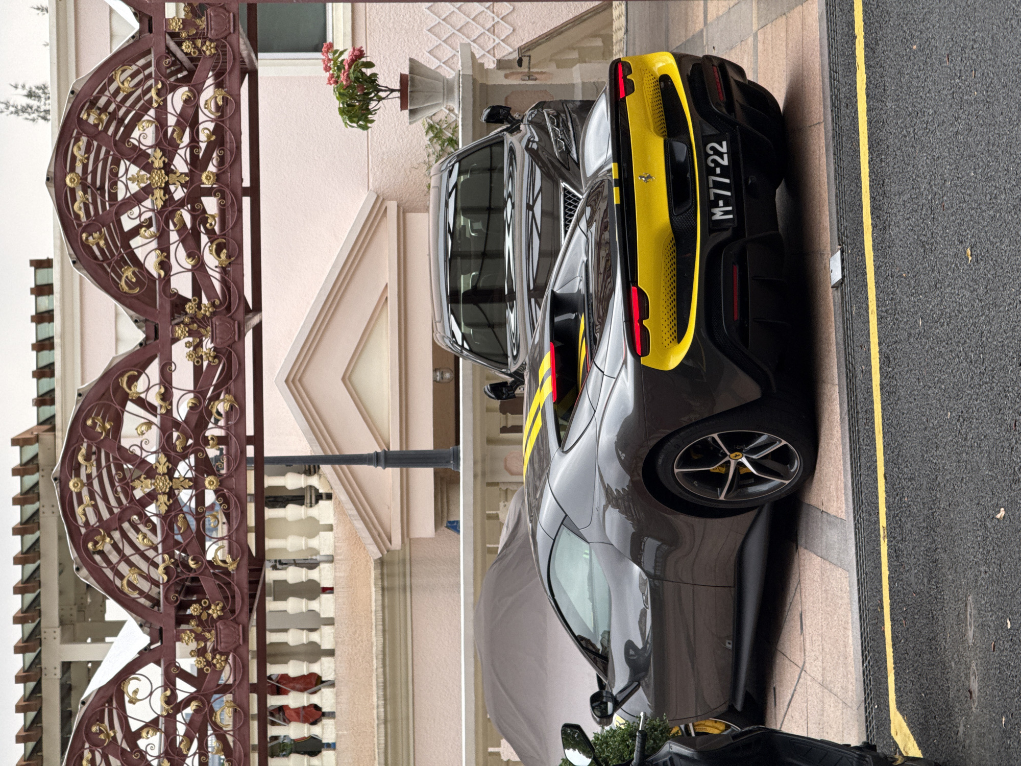 The image shows two cars parked adjacent to a road in Macau, China. The car closest to the viewer is a gray sports car with a yellow stripe and the license plate "M-77-22". Behind it is a gray SUV. The cars are parked in front of a light pink building featuring a gabled entrance and a flower pot on a pedestal. A wrought-iron gate with intricate gold details is partially visible on the left side of the frame. The road is asphalt and has a yellow curb.