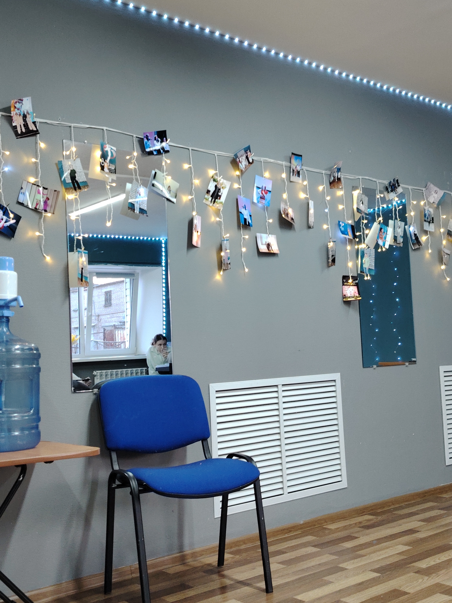A room in Chaikovsky, Russia, features a gray wall adorned with string lights displaying photos. Below, a blue chair sits near a light wood-colored table with a large water bottle. A mirror reflects an individual sitting by a window, while ventilation grates are positioned on the wall. The floor is made of light wood planks.