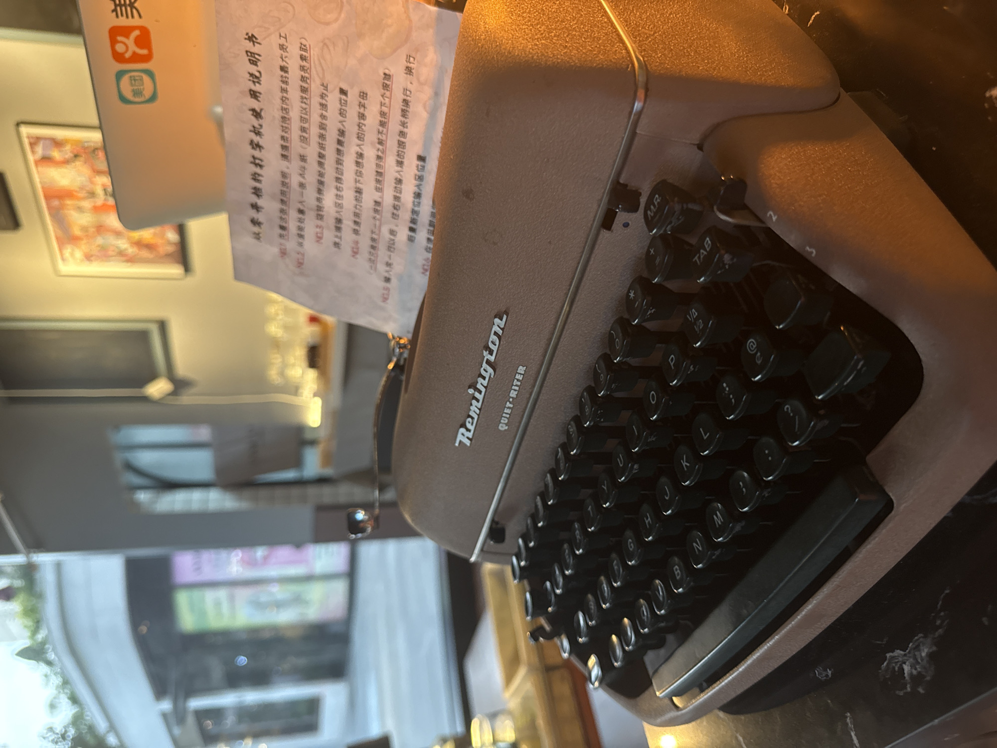 The image shows a close-up of a vintage Remington Quiet-Riter typewriter, positioned on a dark, reflective surface, possibly a marble countertop. The typewriter is a light brown color with black keys. In the background, a white laptop with a logo that appears to be from Meituan (a Chinese e-commerce platform) is visible, along with a framed picture on the wall. A piece of paper with Chinese text, seemingly instructions for the typewriter, is placed on top of the typewriter. The setting appears to be indoors, possibly a cafe or a shop, with blurred background elements suggesting a commercial environment. The lighting is warm and somewhat dim, casting shadows on the objects. The overall scene suggests a display or a functional setup for using the typewriter, potentially in Yiwu, China, given the presence of the Meituan logo. There are no people visible in the frame.