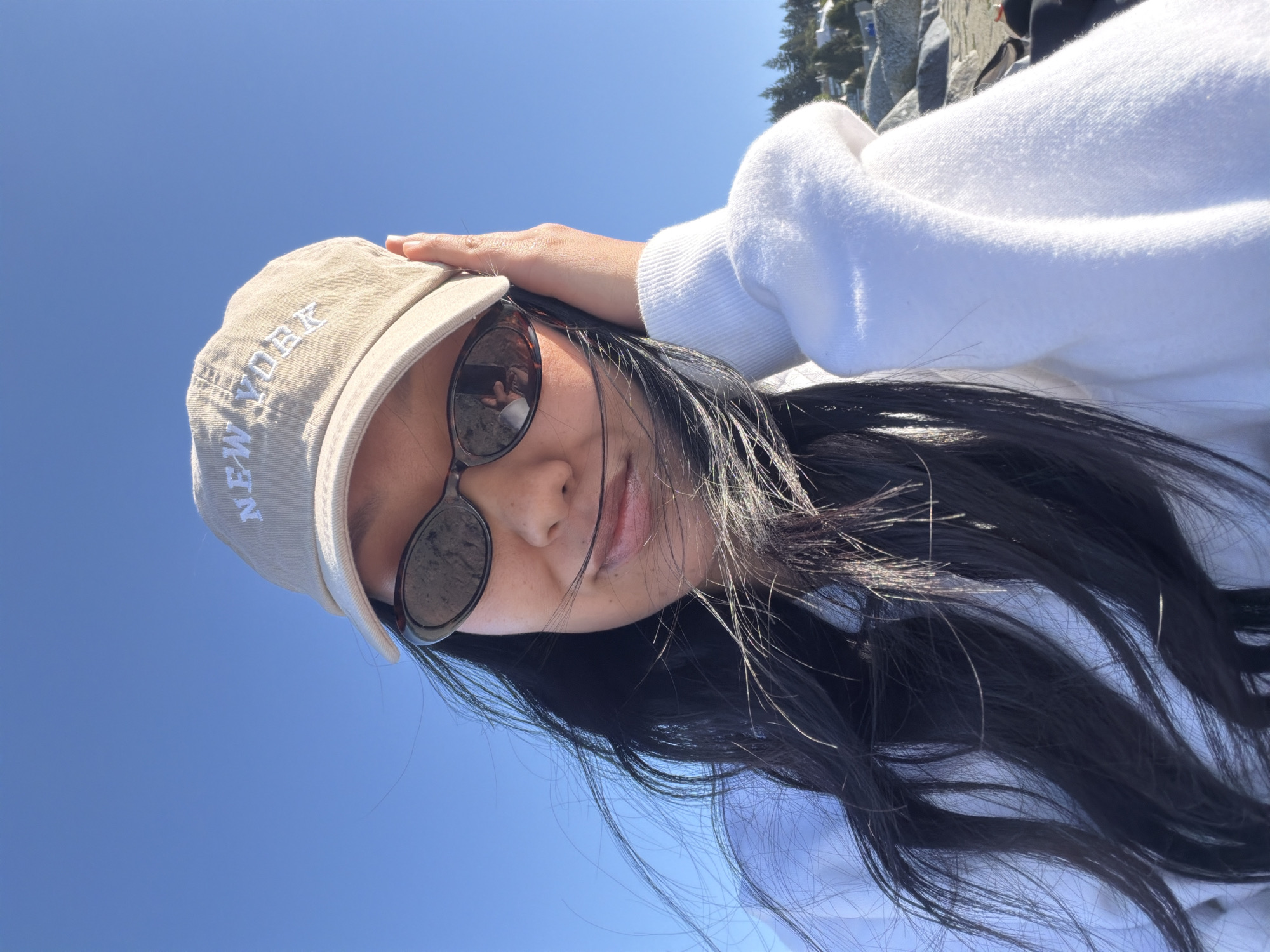 A medium-shot angled portrait depicts a young woman. She is wearing a tan baseball cap with "NEW YORK" stitched in white across the front. She is also wearing black sunglasses, under which her dark hair cascades. She is wearing a white sweatshirt. Her right hand is positioned atop her head, touching the cap.
The setting is outdoors, with a bright blue sky serving as the backdrop. A few indistinct structures, potentially rocky outcroppings or buildings, are visible at the top of the frame. The specific location in Vancouver, Canada is unidentifiable.