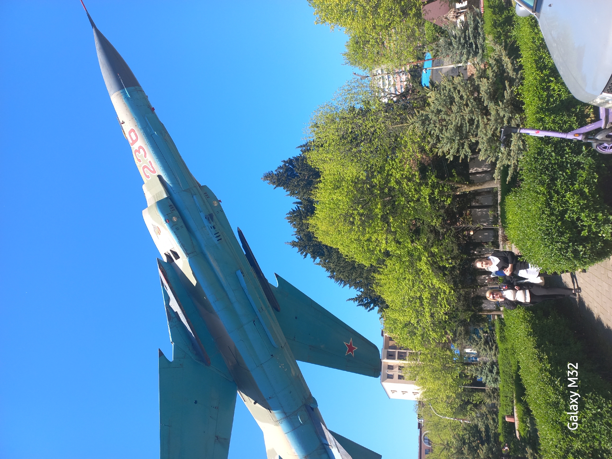 The image captures an upward-angle view of a MiG-21 fighter jet on display in Sochi, Russia. The aircraft, painted in a faded blue, is adorned with the number "230" in red lettering on its nose. The tail fin features a red star emblem. A clear blue sky serves as the backdrop for the jet.
To the right, the scene transitions to a park setting. Green trees and trimmed hedges are visible. Two women are standing on a paved walkway. One is wearing a white and black school uniform. A scooter is nearby.