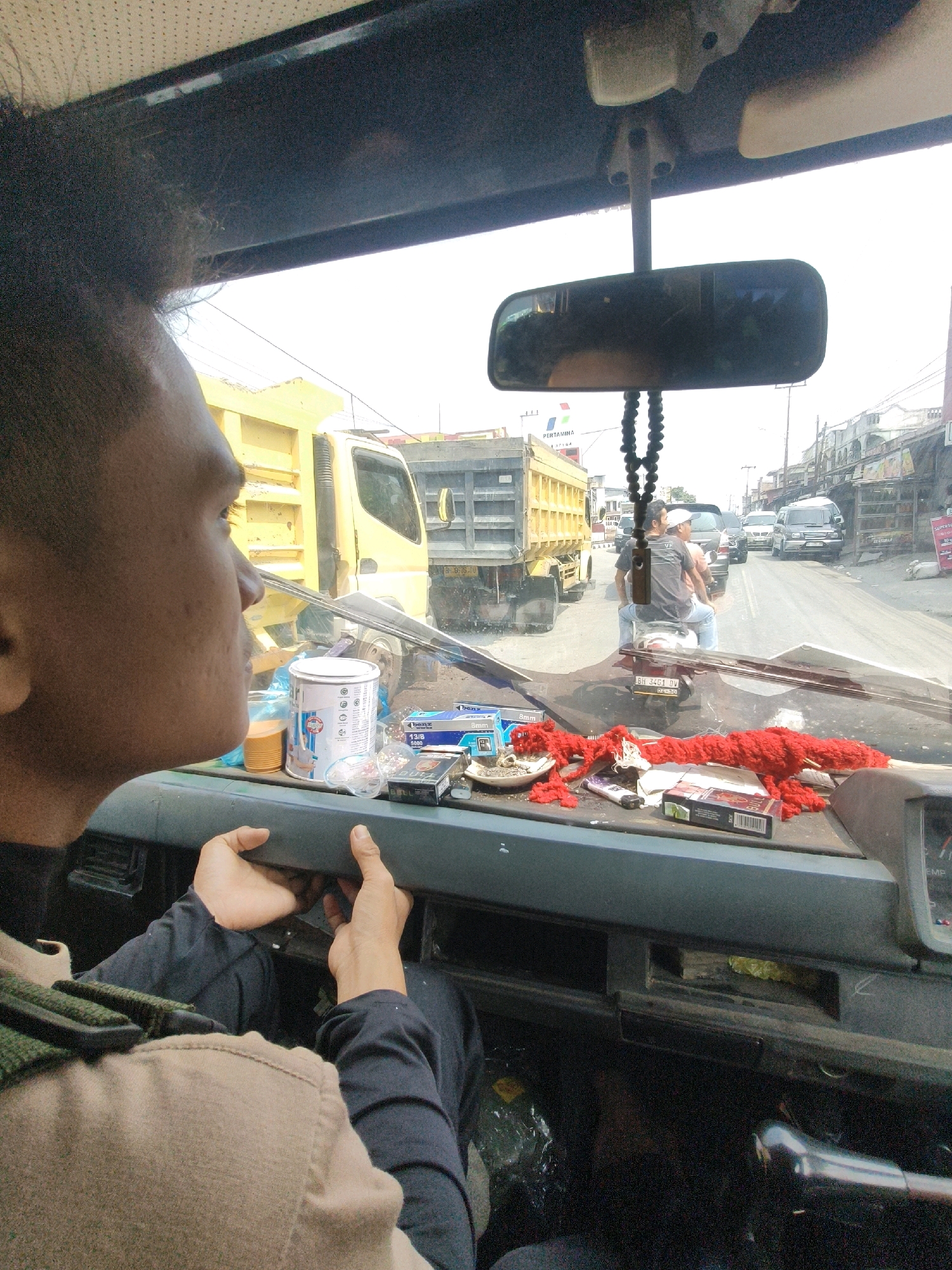 The image is taken from inside the cabin of a truck, looking out through the windshield. A person, likely the driver, is visible in the foreground, their face turned to the left. Their hands are on the dashboard, appearing to adjust something. The dashboard itself is cluttered with various items including cigarette packs, a can, a small plate with what appears to be a cigarette butt, and a red fuzzy object. Outside the truck, traffic is visible on a paved road. There are other vehicles, including a yellow dump truck and a motorcycle carrying two people. Buildings line the side of the road, suggesting an urban or semi-urban environment. The sky is overcast. The location is identified as the city of Lubuk Nagodang, Indonesia, implying a Southeast Asian setting with local architecture and vehicle types.