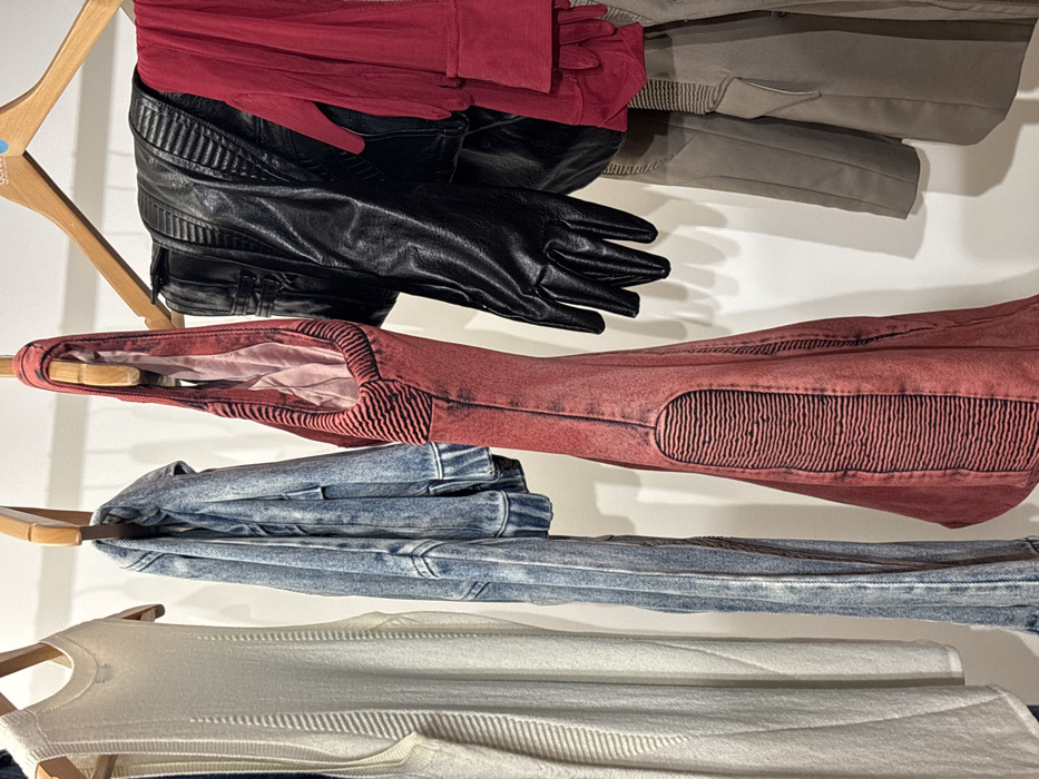 The scene depicts an indoor environment, likely a retail space or wardrobe, with various clothing items hanging on wooden hangers against a plain white wall. From left to right, the objects include: a cream or off-white sleeveless knit garment with vertical ribbed texturing; a light blue acid-wash denim jacket; a reddish-pink distressed fabric garment, possibly a dress or long top, featuring black horizontal ribbed patterns on its front panels; a black faux-leather jacket with ribbed shoulder details, accompanied by a pair of black faux-leather gloves hanging beneath it; and a vibrant red sheer fabric garment, partially visible, hanging above a beige or khaki-colored jacket. All garments are suspended from wooden hangers. A blue label, partially visible at the top right, shows the letters "GETD". The specific location within London, United Kingdom, is not discernible from the provided image.