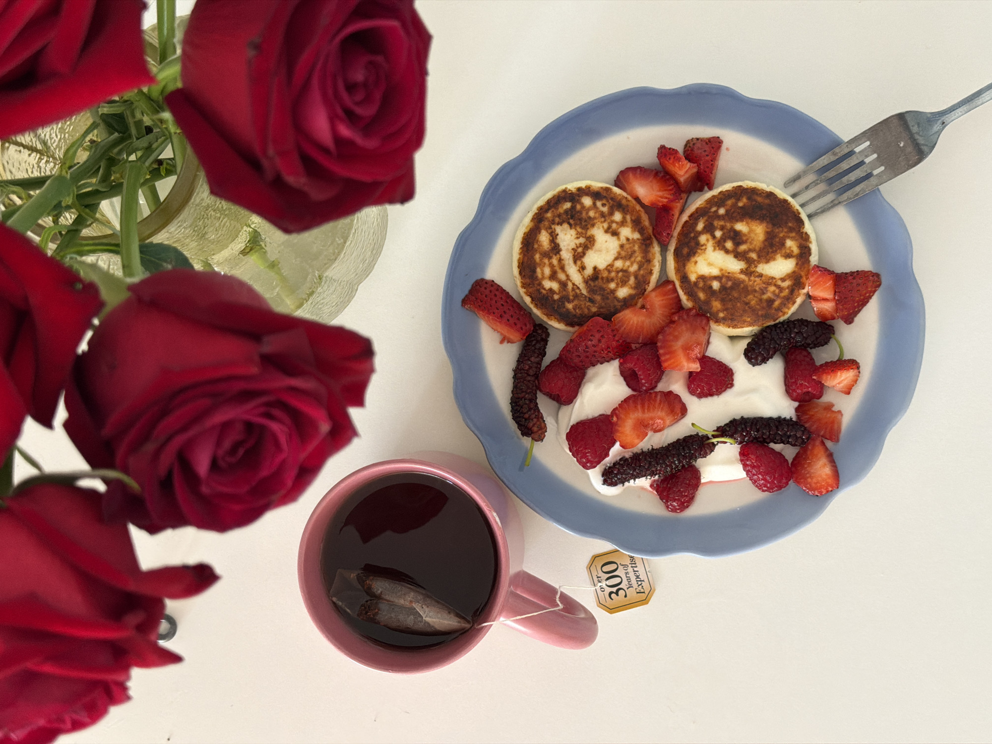 The image presents a still life composition. On a white surface, a bouquet of red roses is arranged in a clear glass vase. Adjacent to the roses, a light blue plate with a ruffled edge holds two golden-brown pancakes, topped with fresh strawberries, raspberries, and mulberries. A dollop of white cream sits beneath the fruit. A silver fork rests on the plate. Next to the plate, a pink mug contains a dark liquid, presumably tea, with a tea bag steeping inside. The tea bag’s tag is visible, bearing the words "300 Years of Expertise." There is no evidence to indicate this image was taken in Tel-Aviv, Israel.