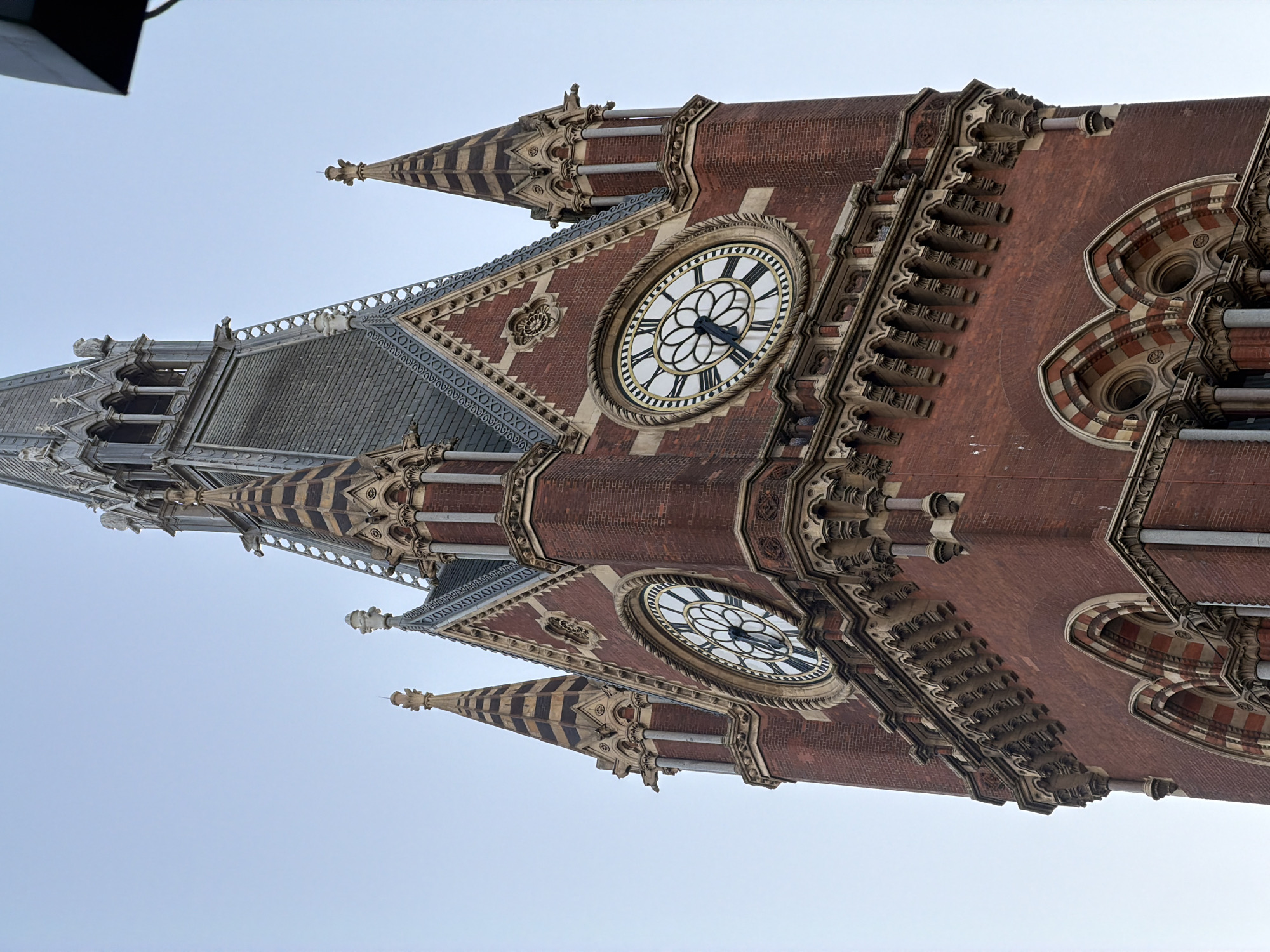 The image presents a striking low-angle view of a prominent section of a grand Victorian Gothic Revival building, likely a clock tower or a major facade, set against a clear, bright blue sky. The structure is characterized by its rich red brickwork complemented by extensive use of light-colored stone for intricate decorative elements, carvings, and architectural details.
Two large, circular clock faces are visible on the red brick walls. The upper clock, more fully visible, features black Roman numerals and hands, indicating the time to be approximately 11:20. Its face also includes an ornate, floral-like pattern at its center. The lower clock face, partially obscured, appears to share a similar design.
The building boasts multiple pointed spires and turrets, some adorned with distinctive horizontal black and light-colored striped patterns. Elaborate stone ornamentation, including decorative railings, tracery, and gargoyle-like figures, frames windows and cornices, adding to the building's intricate detailing. Portions of a dark, slate-grey roof are also visible.
The perspective is from below, looking sharply upwards, which emphasizes the towering scale and intricate craftsmanship of the architecture. The clear sky suggests daytime and fair weather conditions. Based on its distinctive architectural style and the provided context, the building is identifiable as St Pancras International in London, United Kingdom. No people or specific activities are depicted, with the focus solely on the magnificent structure itself.