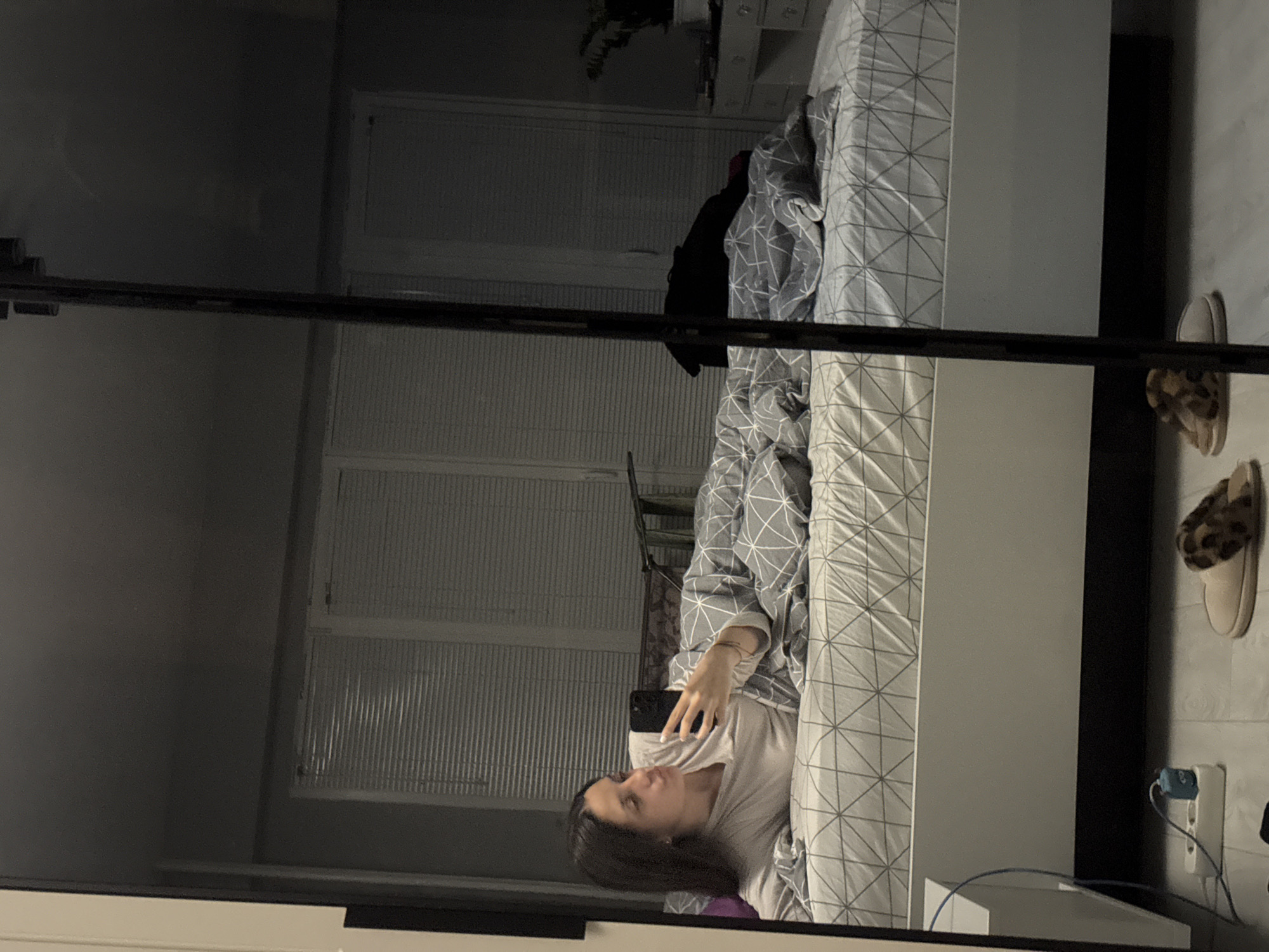 A woman is lying on a bed in a room that is reflected in a mirror. The woman is holding a phone. The bed has a gray and white patterned comforter. The room has white window blinds. The electrical outlet is mounted on the wall with a plug in it. A pair of slippers are on the floor next to the bed. The image was taken in Sofia, Bulgaria.