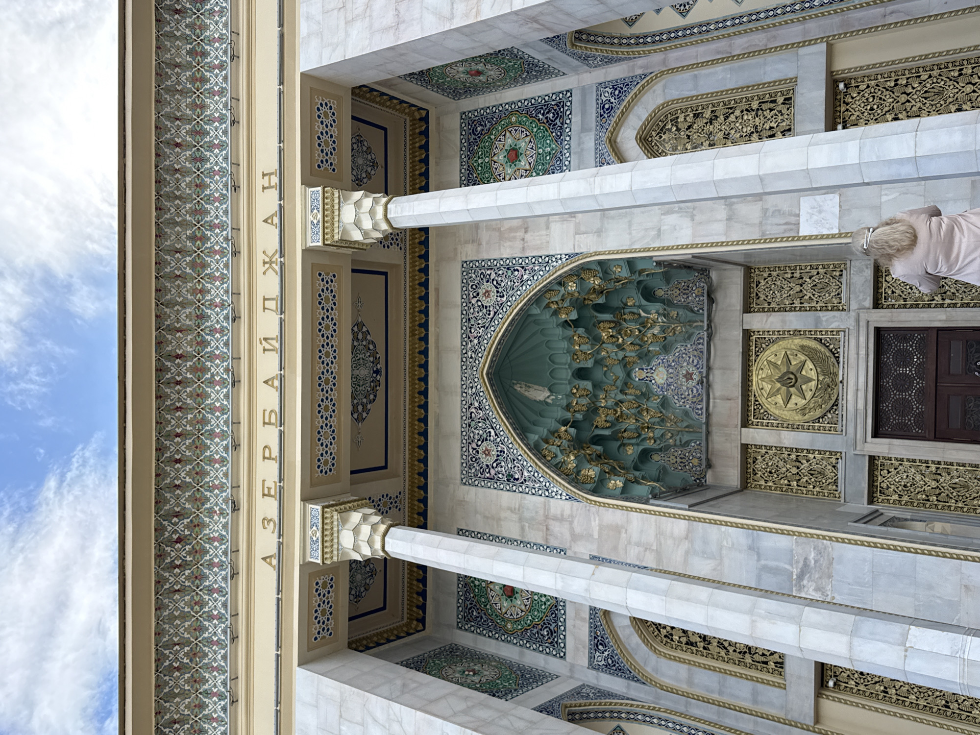 This media file captures an interior view of the Moscow Cathedral Mosque in Moscow, Russia, likely during the daytime given the bright sky visible on the left. The camera is angled upwards, showcasing the intricate Islamic architectural details of the mosque's ceiling and upper walls. The scene is dominated by elaborate geometric patterns, vibrant tilework in shades of blue, green, and white, and decorative golden friezes. Large, fluted marble columns rise towards the ceiling, supporting arched structures adorned with ornate carvings.
A prominent feature is a large, teal-colored niche with golden relief work depicting what appears to be branches and leaves. Below this niche, a circular golden emblem with star-like points is visible. On the far right, a dark wooden door with intricate latticework can be seen, suggesting an entrance or a separate chamber. A woman with blonde hair, wearing a light pink or beige garment, is partially visible on the right side of the frame, positioned as if observing or praying. The overall impression is one of grandeur, artistry, and spiritual serenity.
Vertical text in Cyrillic script is present on a decorative panel running along the left side of the image, which reads "АЗЕРБАЙДЖАН" (Azerbaijan). This likely indicates a connection to Azerbaijan, perhaps through sponsorship or the origin of the architectural inspiration. The weather appears clear with blue skies and scattered white clouds, visible through an opening on the far left, suggesting fair weather.