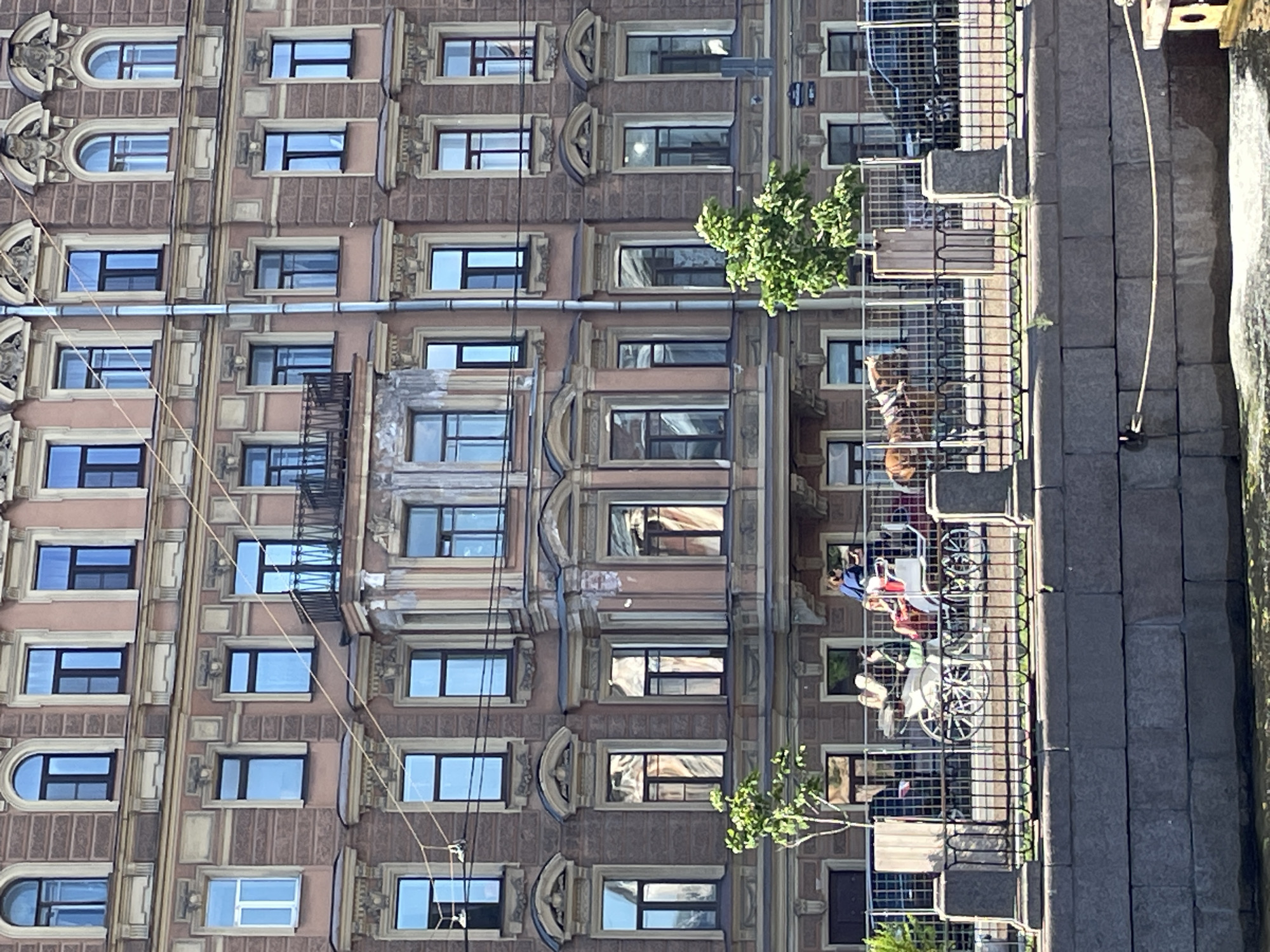 The image depicts a street scene in Saint Petersburg, Russia, with a focus on a multi-story building and a cobblestone walkway. The building's facade is adorned with architectural details, including decorative moldings around numerous windows. The windows themselves vary in design, with some being rectangular and others arched. Wires are strung across the building, suggesting an urban environment.
On the walkway, several people are visible, some seated in what appears to be a horse-drawn carriage. The carriage has large spoked wheels and is occupied by at least two individuals. Another person stands near the carriage. The walkway is lined with a metal fence, behind which the building stands. Beyond the walkway, a body of water, possibly a canal or river, is visible, with some ripples suggesting movement. The light and shadows suggest it is daytime, likely sunny.
The scene appears to be a tourist area, with the carriage likely offering rides. The architecture and the presence of a waterway are characteristic of Saint Petersburg. No visible text is present in the image.