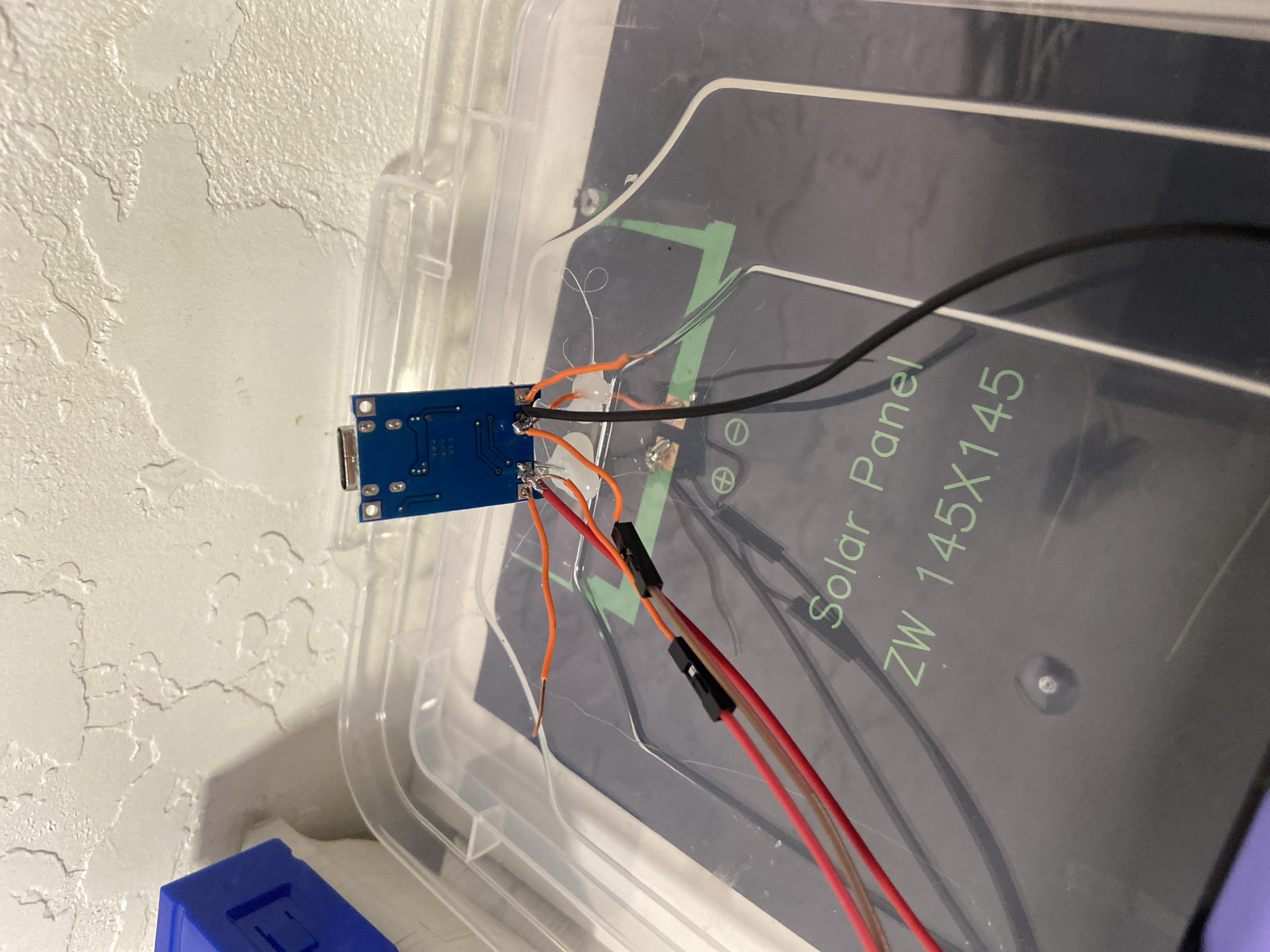 In a close-up, overhead shot, a blue circuit board is connected to wires. The circuit board is positioned on top of a black solar panel labeled "Solar Panel ZW 145X145." Several wires, in red, orange, and black, are soldered to the circuit board and extend downwards towards the solar panel. Some of the wires are connected to black plastic connectors. The solar panel itself is partially visible within a clear plastic container. The background consists of a textured, light-colored wall. The scene is indoors, with no discernible urban or environmental context.
