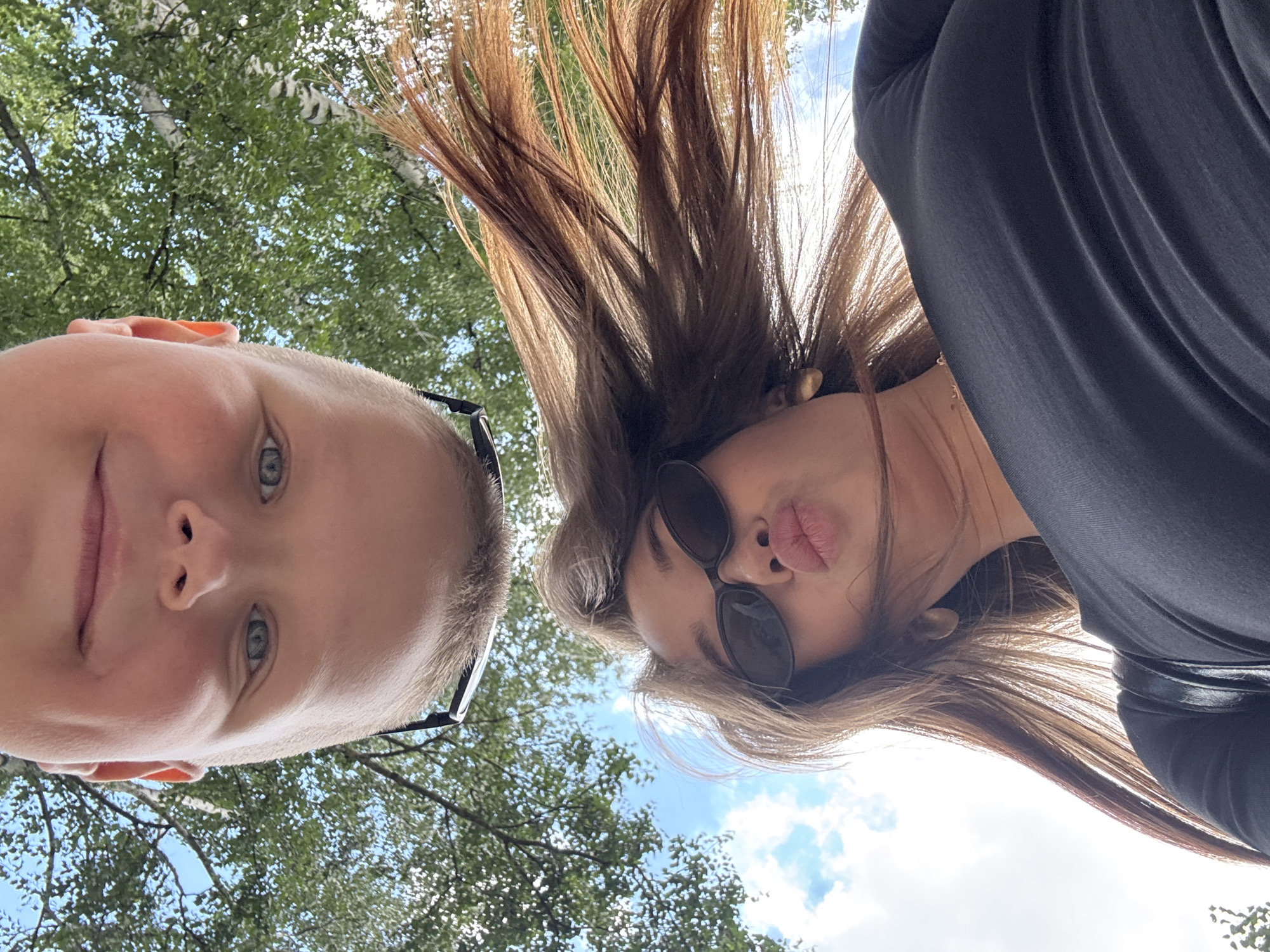 An inverted, low-angle outdoor shot features two individuals. At the top of the frame, a person's face, viewed upside down, shows blue eyes, a nose, and a mouth with lips pursed. The person appears to be wearing glasses. Below, another person, also upside down, is visible from the shoulders up. This individual has long, brown hair that is windswept, brown eyes, and is wearing black sunglasses and a black top. Their lips are also pursed. The background consists of a bright blue sky with scattered white clouds and lush green foliage from trees, suggesting an outdoor, possibly park-like setting. The image was taken in the city of Chaikovsky, Russia.