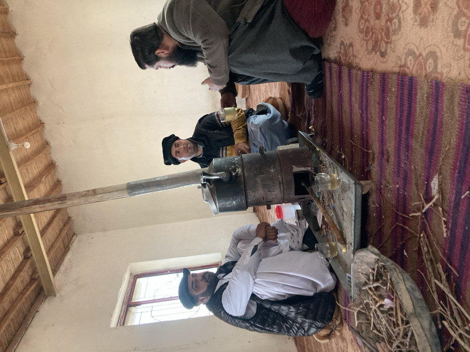 Inside a room in Sanjawi, Pakistan, three adult males are seated on a carpeted floor. A central, dark-colored cylindrical metal stove with an upward-extending chimney is active. A metal cooking pot with a lid rests on top of the stove. The stove is positioned on a rectangular metal tray which also holds two clear glasses containing a light-colored liquid, along with scattered wood fragments. To the left of the stove tray is a boat-shaped basket filled with small twigs and branches.
The man on the left is seated cross-legged, wearing a white traditional garment, a dark quilted sleeveless vest, and a dark flat cap. His hands are clasped in front of him.
The man in the center is seated cross-legged, wearing a striped sweater under a dark sleeveless vest, light-colored trousers, and a dark Pakol-style cap. He holds a clear glass with a light-colored liquid in his right hand.
The man on the right is seated cross-legged on a maroon cushion, wearing a dark, long-sleeved garment and dark trousers. He possesses a prominent dark beard and mustache. He holds a clear glass with a light-colored liquid in his left hand and is observed gesturing with his left hand while facing the central man.
The room features light-colored walls, a window with a dark frame and metal security bars on the left, and a ceiling constructed with exposed wooden beams and slats. A single bare light bulb hangs from the ceiling. The floor is covered with a striped purple and brown rug transitioning to a patterned beige rug. Scattered debris, including small wood fragments, is visible on the floor around the stove. A small, red, cylindrical plastic container is positioned behind the stove.