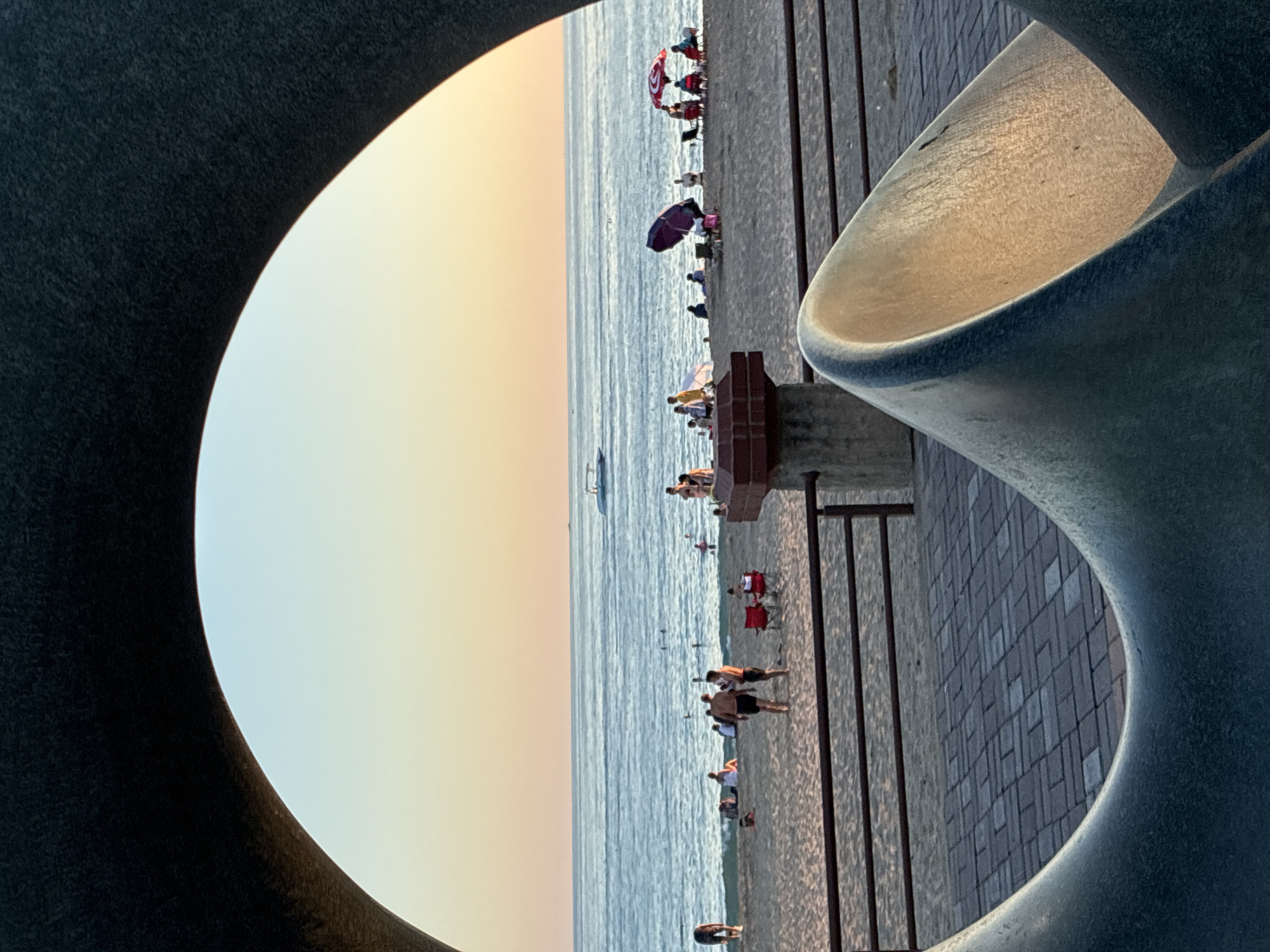 This photograph captures a view of the beach in Alanya, Turkey, framed by a large, dark, circular sculpture in the foreground. The sky transitions from a pale orange at the horizon to a clear light blue. The sea is a deep blue with gentle waves and is populated by several small boats and a few people swimming.
On the sandy beach, which is lined with a metal railing and a brick walkway, numerous people are engaged in various beach activities. Some are sunbathing, while others sit in beach chairs or under umbrellas. There are two red beach chairs placed near the water, and a purple umbrella and a red and white umbrella are also visible, providing shade.
In the mid-ground, a concrete structure with a reddish-brown top stands near the railing. Two men, appearing to be in their late 20s or 30s, are standing on the sand, facing each other and seemingly engaged in conversation. One is wearing black swim trunks, and the other is wearing dark swim trunks. To their left, a person is bent over, possibly adjusting their footwear. Further down the beach, more individuals are scattered, some sitting and others walking.
The overall impression is a relaxed, late afternoon scene at a popular beach destination. The sculpture in the foreground adds an artistic and framing element to the otherwise natural beach vista.