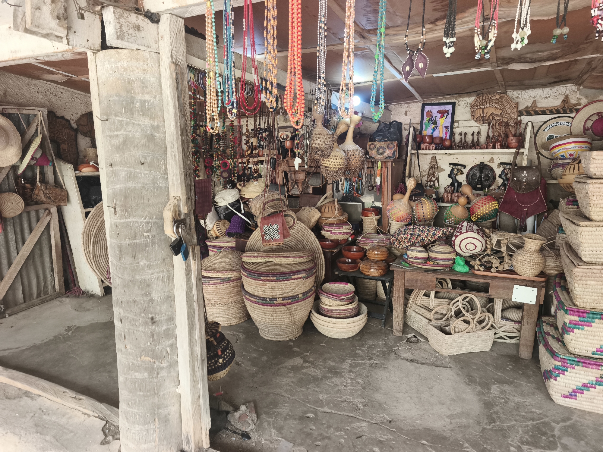The image shows the interior of a small shop in Jos, Nigeria. The shop sells handmade goods, including woven baskets, necklaces, carvings, and bags. The goods are displayed on shelves, tables, and hanging from the ceiling. The floor is concrete and slightly dirty. There are no people visible in the image. The entrance of the shop is supported by a rough-hewn post, seemingly a repurposed palm trunk.