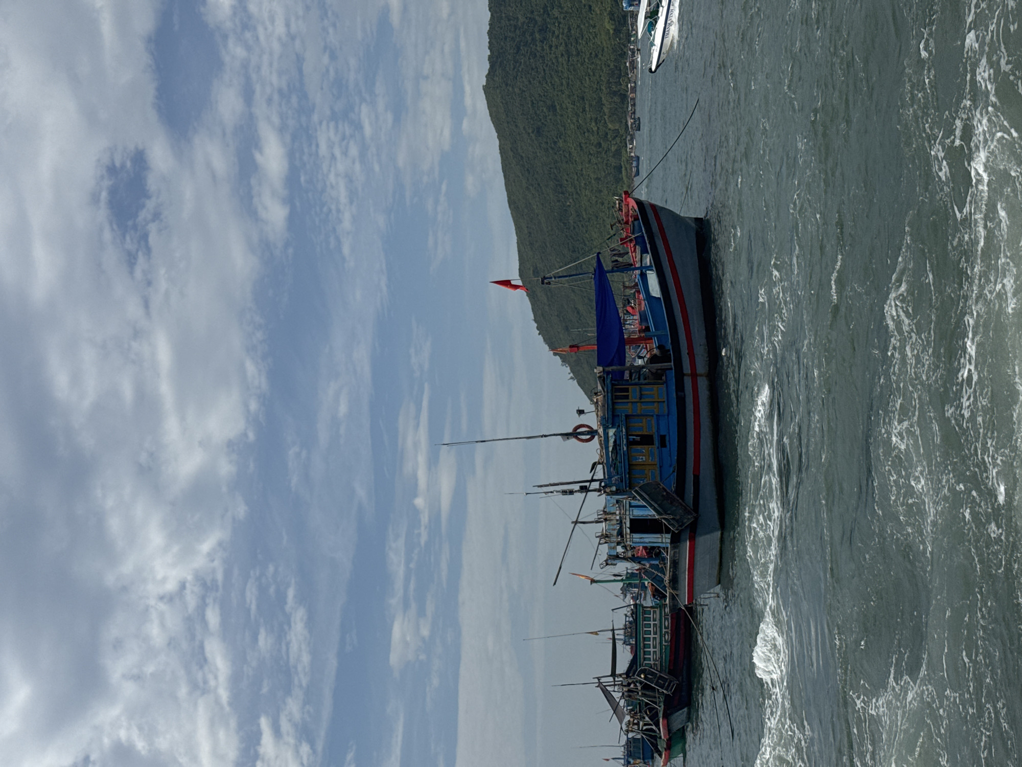 In Nha Trang, Vietnam, several fishing boats are moored in the ocean under a cloudy sky. The water is choppy, with white foam suggesting recent movement. In the foreground, the bow of a blue and red fishing boat is prominent. Further back, a line of fishing boats is visible, with some appearing older and weathered. A lush, green, forested hill with a small village at its base forms the background. To the right, a modern white speed boat is partially visible, with its name, "THANH VINH," legible. The scene depicts a working harbor or bay, common in coastal Vietnamese cities.