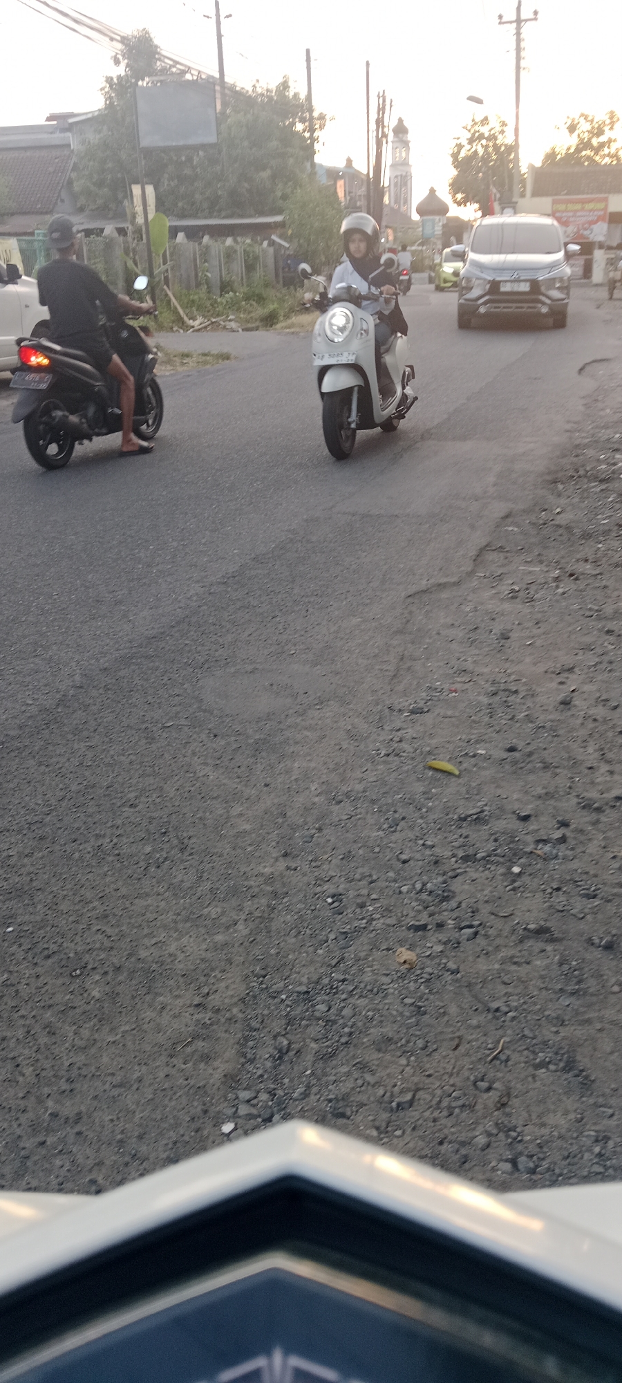 An eye-level, medium shot shows two motorcycles and one car on a paved road in Maguwoharjo, Indonesia. The sun is setting, casting a warm glow over the scene and creating long shadows. The road is asphalt with some gravel and cracks. On the left, a black motorcycle is parked with its brake light illuminated. A person wearing a black t-shirt and a black baseball cap is seated on the motorcycle, facing left. On the right, a white scooter is moving forward, with a person wearing a light blue shirt and a helmet riding it. The person on the scooter is looking forward. In the background, a silver SUV is moving away from the viewer. Behind the vehicles, there are trees, buildings, and utility poles. The sky is a pale orange, indicating dusk.
