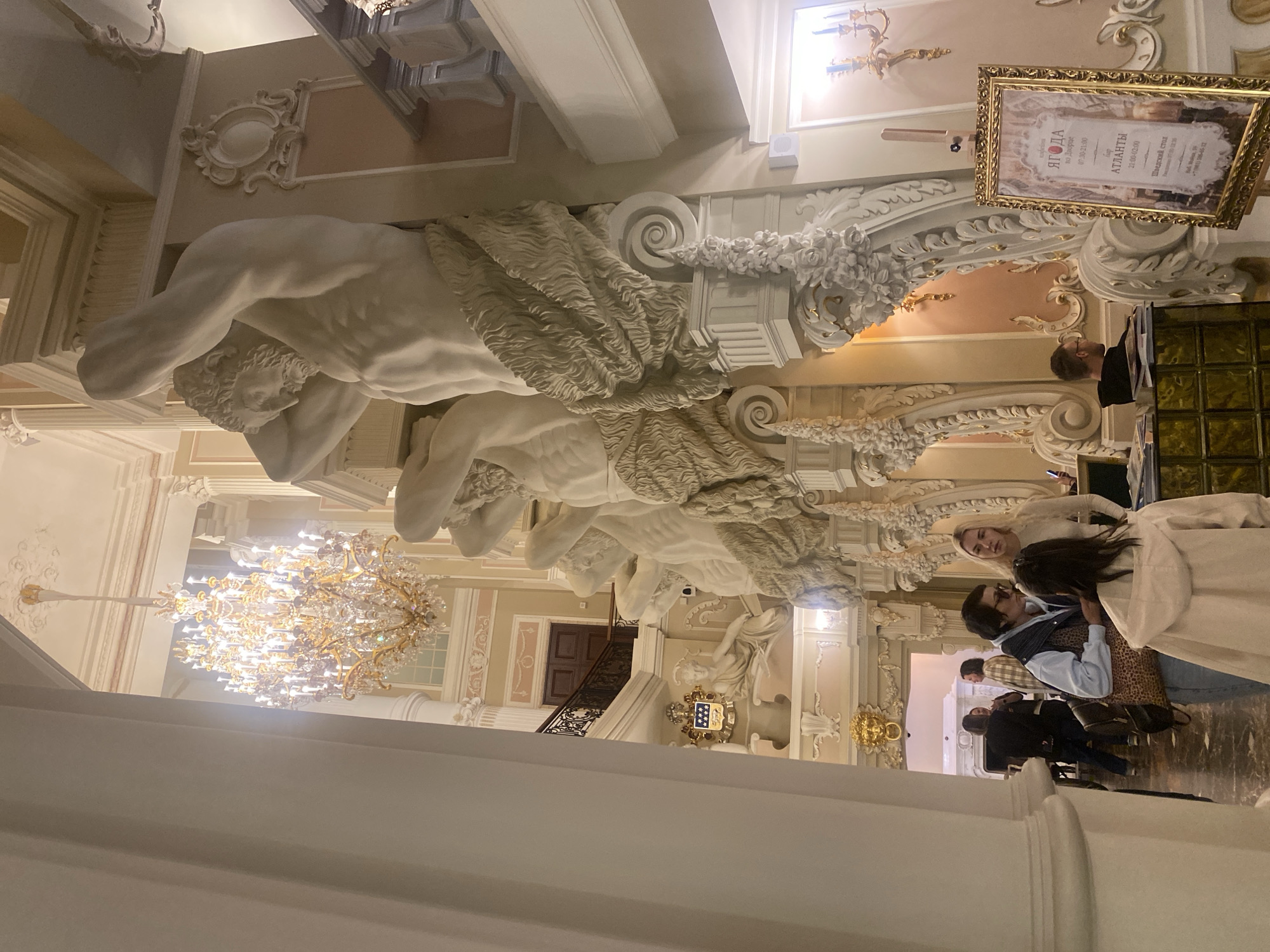 The image shows a grand interior with ornate architecture and a large chandelier. In the foreground, several people are gathered, some conversing. To the right, a framed poster is displayed on an easel. The poster features Cyrillic text and appears to be an advertisement for a venue or event. The overall impression is one of luxury and sophistication. Based on the poster's text, which includes "Шведский стол" (Swedish table, or buffet) and an address that likely translates to Nevsky Prospekt, and the style of the interior, this scene is likely located at the Astoria Hotel in Saint Petersburg, Russia, a historic hotel known for its opulent decor and central location.