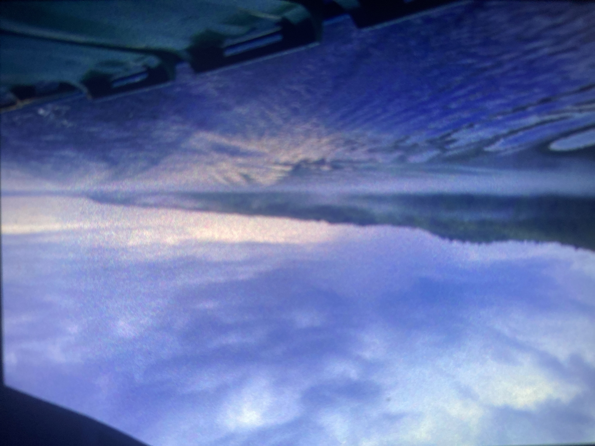 The image displays a tranquil landscape scene, inverted vertically from a conventional perspective, featuring a sky above a body of water with a distant shoreline.
The upper portion of the image (which would be the sky if rotated 180 degrees) is a vast expanse of sky. It is dominated by soft, diffused clouds in a blend of serene blue, purple, and white hues. Lighter patches of pink or faint yellow suggest the soft light of either dawn or dusk.
A distinct, slightly irregular dark band stretches horizontally across the mid-section, forming a horizon line that separates the sky from the water. This band appears to be a distant, low-lying landmass, possibly a forested shoreline or hills, which is somewhat blurred by distance and atmospheric conditions. Above this landmass, there's a brighter, almost luminous band of light, hinting at the sun's position just at or below the horizon.
The lower portion of the image (the water if rotated 180 degrees) shows a calm body of water, likely a lake or a large river. It effectively reflects the colors and soft light of the sky above, creating an almost symmetrical composition. Gentle ripples are visible on the water's surface, subtly distorting the reflected light and colors. Towards the very bottom edge of the frame, dark, indistinct shapes are visible, which are reflections of objects positioned above the water, such as parts of a boat, a dock, or an overhead structure.
No people or specific activities are visible, emphasizing the quiet, natural environment. The overall atmosphere is peaceful and serene. The time of day is clearly either sunrise or sunset, given the soft, pastel colors and diffused light. There are no specific visual cues within the image that explicitly confirm the location as Saint Petersburg, Russia; it depicts a general natural landscape. The image has a slightly grainy quality. No text is visible.