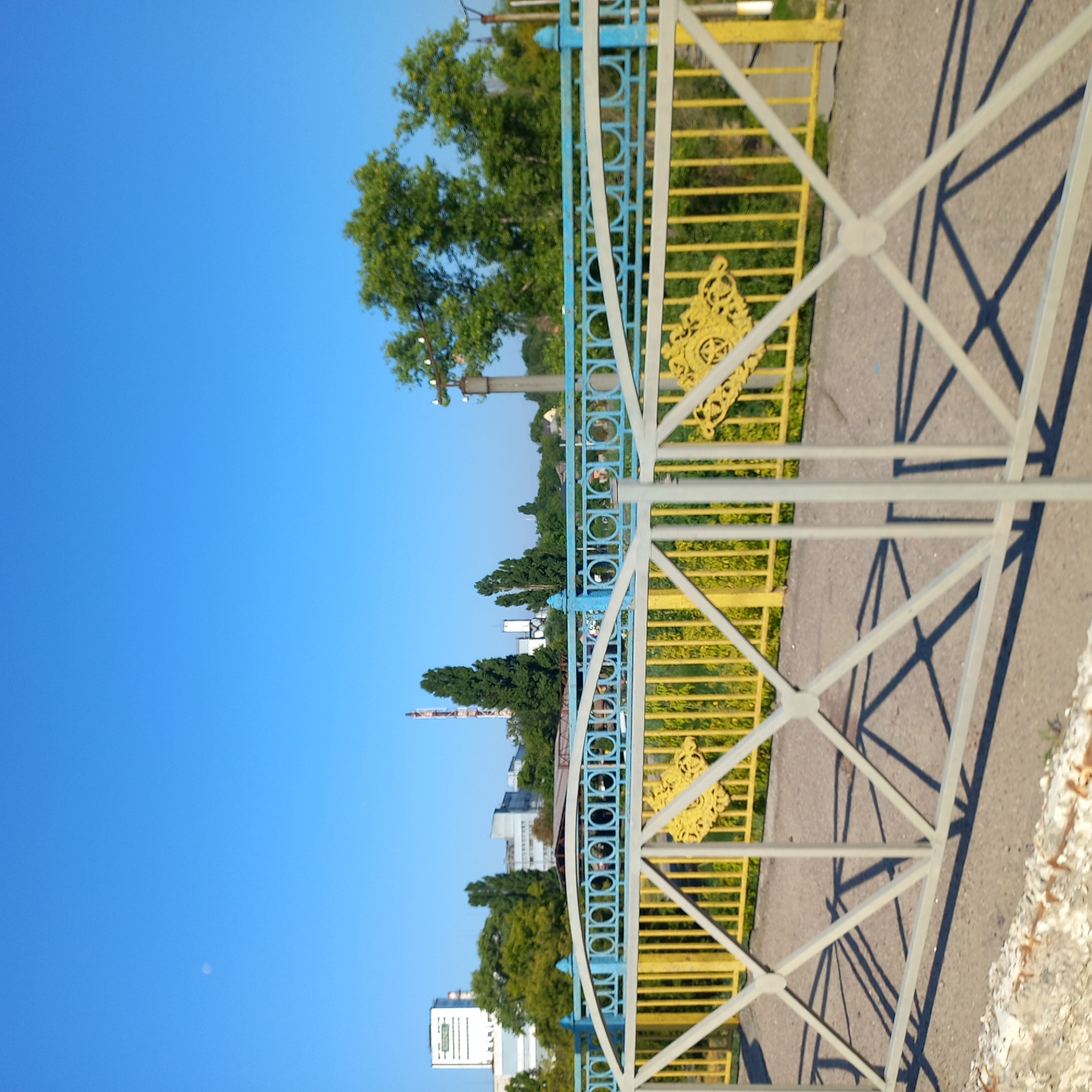 The image shows a view of industrial buildings and green trees under a clear blue sky with a visible moon. In the foreground, there is a metal fence with a grey outer railing and yellow and blue inner railings, decorated with ornate yellow accents. A paved walkway runs behind the fence. In the background, several industrial structures, including a tall white building and a red and white smokestack, are visible amidst lush green trees. The scene appears to be a clear, sunny day. This is a view of an industrial area in Kropyvnytskyi, Ukraine, likely near a bridge or pedestrian walkway.