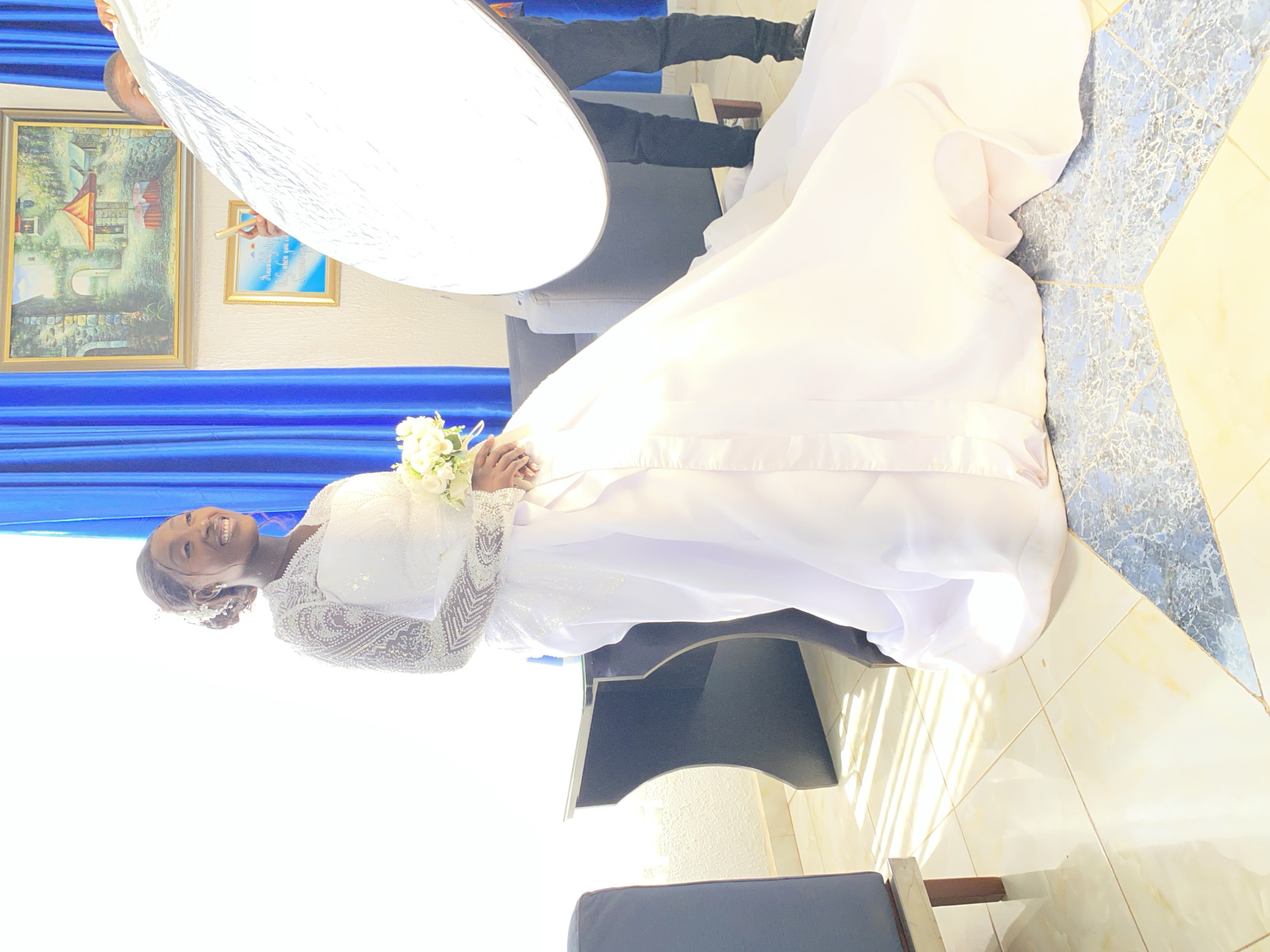 A woman in a white wedding dress smiles and holds a bouquet of flowers. A person holds a light reflector above the woman. The woman is standing on a tile floor. A blue curtain hangs in the background, along with two framed pictures. A gray couch and dark-colored furniture are also visible. The location is likely indoors, potentially in Barakin Angundi, Nigeria.