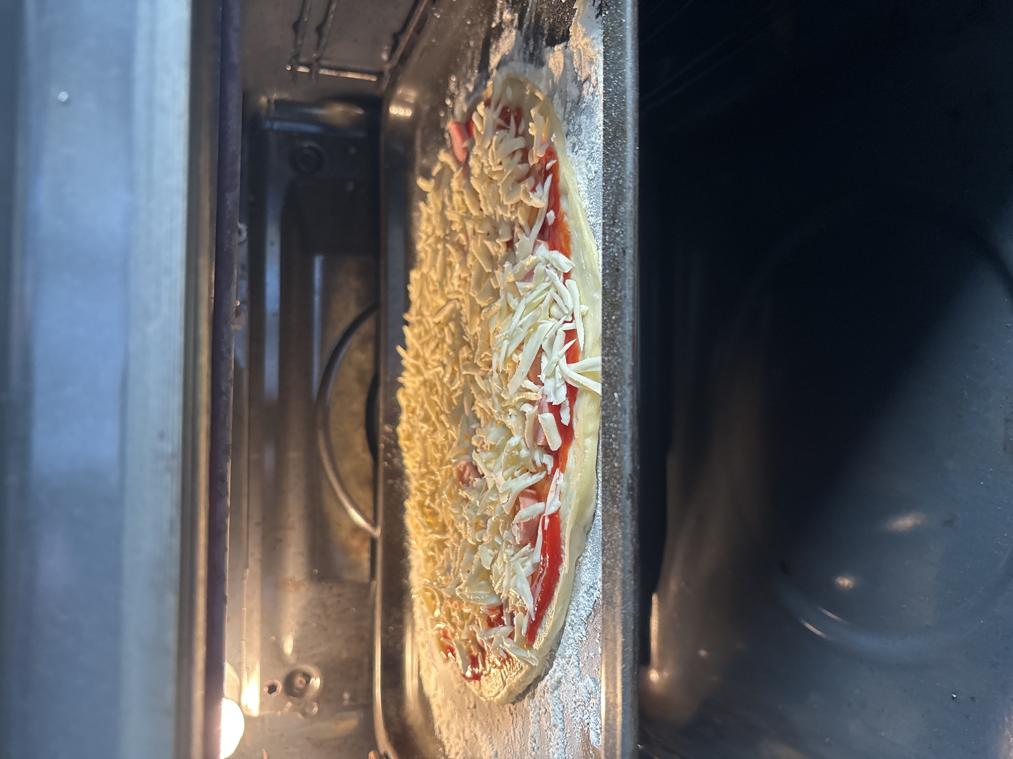 An uncooked pizza is on a baking sheet in an oven. The pizza has a red sauce, some pink toppings that appear to be ham, and shredded white cheese. The baking sheet is dusted with white flour and is resting on the oven rack. The oven interior is visible, including the heating element at the top and the dark, reflective oven floor. The lighting suggests the oven light is on. This scene is not specific to any city or location.
