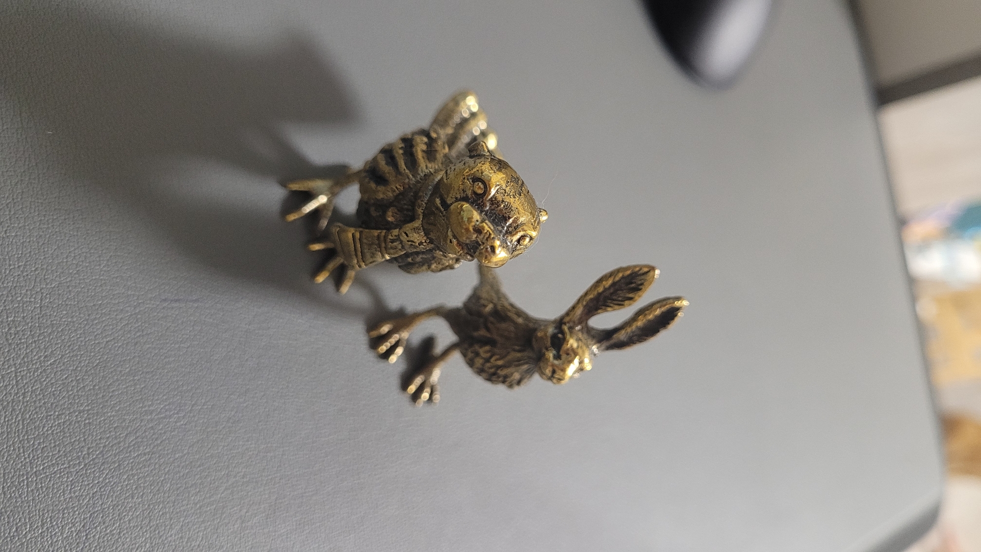Two small, antique-looking brass figurines of animals are positioned on a gray surface. The figurine on the left appears to be a bird, with intricately detailed feathers and a scarf-like accessory around its neck. Its eyes are wide open, and its beak is slightly curved. The figurine on the right is a rabbit, with prominent ears standing upright and a textured body representing fur. Both figurines are facing slightly towards the left. The gray surface is smooth with a subtle texture, and there are faint shadows cast by the figurines. The background is out of focus, showing hints of muted colors.