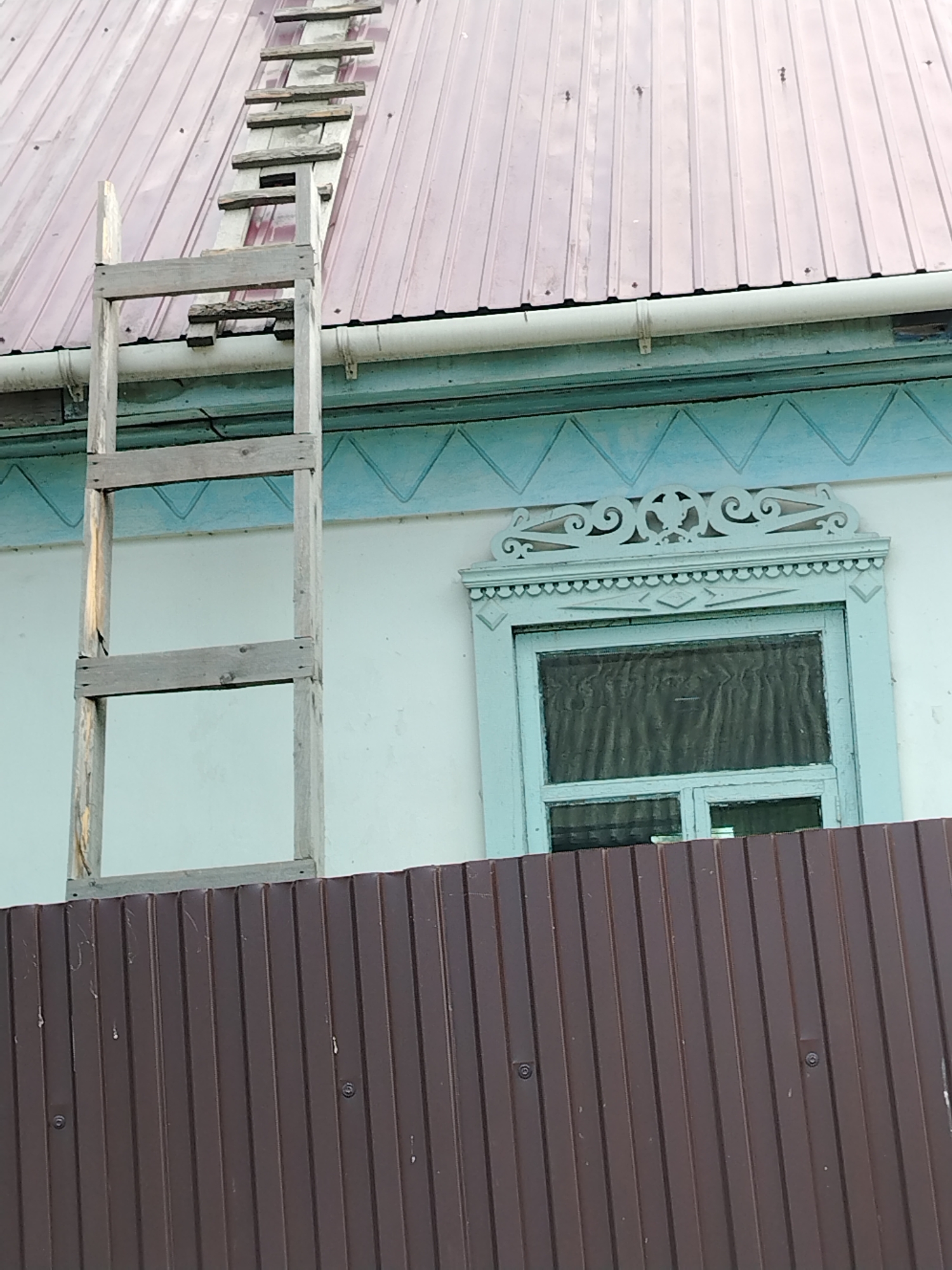 A wooden ladder leans against a reddish-brown metal roof, positioned diagonally across the image. The ladder has visible wooden rungs. Below the roofline, a white wall features decorative blue trim and a window with a light blue frame and a dark curtain. A brown corrugated metal fence runs horizontally across the lower portion of the image, obscuring the base of the wall. The scene appears to be outdoors.
