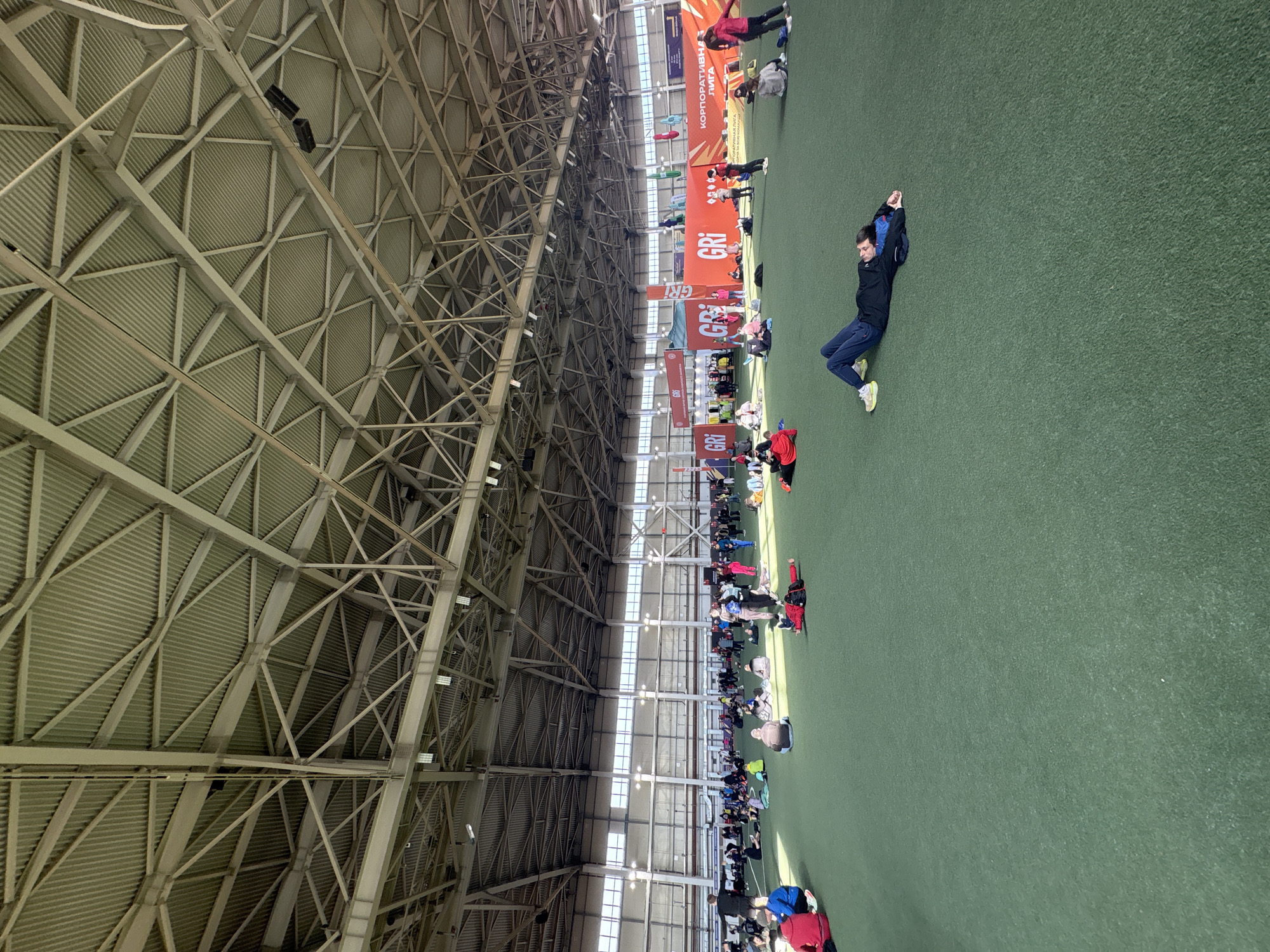 The image shows a large indoor space, possibly a sports arena or exhibition hall, in Matiushino, Russia. The roof structure is a complex network of metal beams. A large group of people is gathered on a green surface, which could be artificial turf or a carpet. The people are arranged in rows and appear to be seated or lying down. Banners with the letters "GRI" are suspended above the crowd. One individual is lying on the turf, with his hands behind his head, and is wearing a dark jacket, blue pants, and light-colored sneakers. The space is well-lit by natural light coming through a row of windows along one side of the structure.