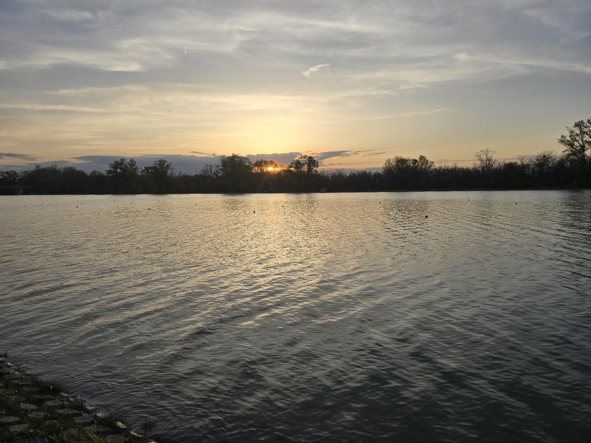 The image depicts a body of water, likely a lake, at sunset or sunrise. The sun is partially obscured by trees on the far bank, creating a bright glare. The water surface is rippled, reflecting the light and creating a shimmering effect. The shoreline is lined with trees and some vegetation. The sky above is a mix of gray and orange hues. There are floating objects visible on the water. The location is Unknown, Unknown.