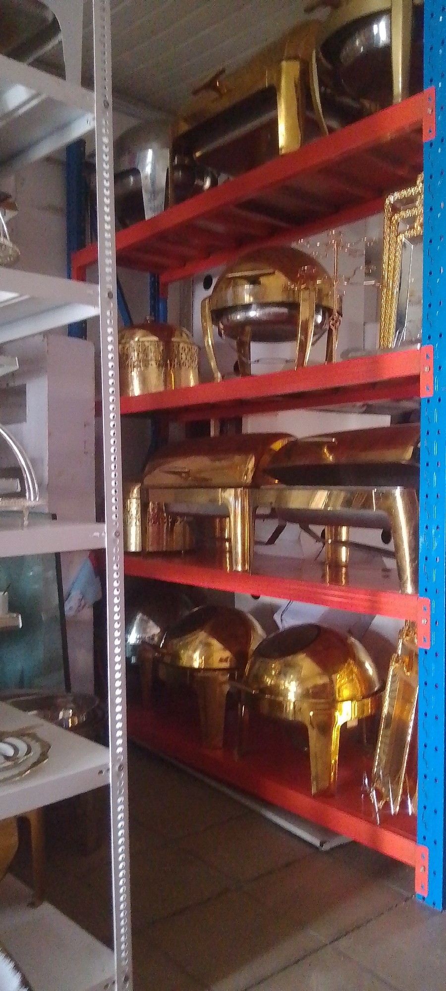 A storage area in Unknown, Unknown, features rows of metal shelving containing various sizes of golden chafing dishes. The shelving units are constructed with red and blue vertical supports, and the shelves themselves are red. The chafing dishes appear to be made of polished metal and are arranged on each shelf. Some of the dishes have lids and others are open. Additional silver and white shelving units are visible on the left side of the frame.