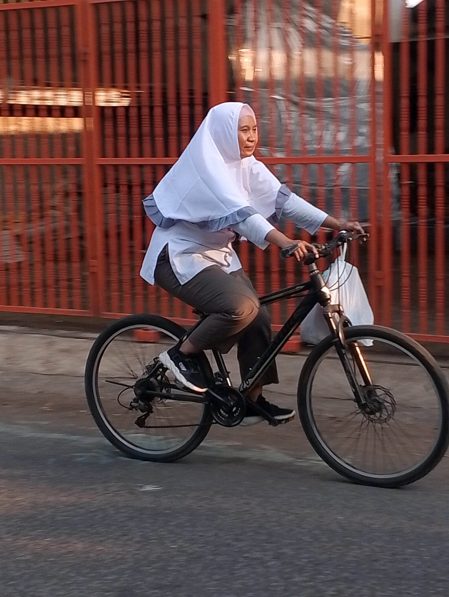 In Probolinggo, Indonesia, a woman wearing a white hijab and a long-sleeved white shirt with grey trim rides a black bicycle. She is wearing dark pants and black athletic shoes. Her right hand is on the handlebars, and a white plastic bag hangs from the handlebars. The bicycle has front and rear suspension and disc brakes. The background features a bright orange metal fence and a blurred asphalt road. The lighting suggests it is either early morning or late afternoon.