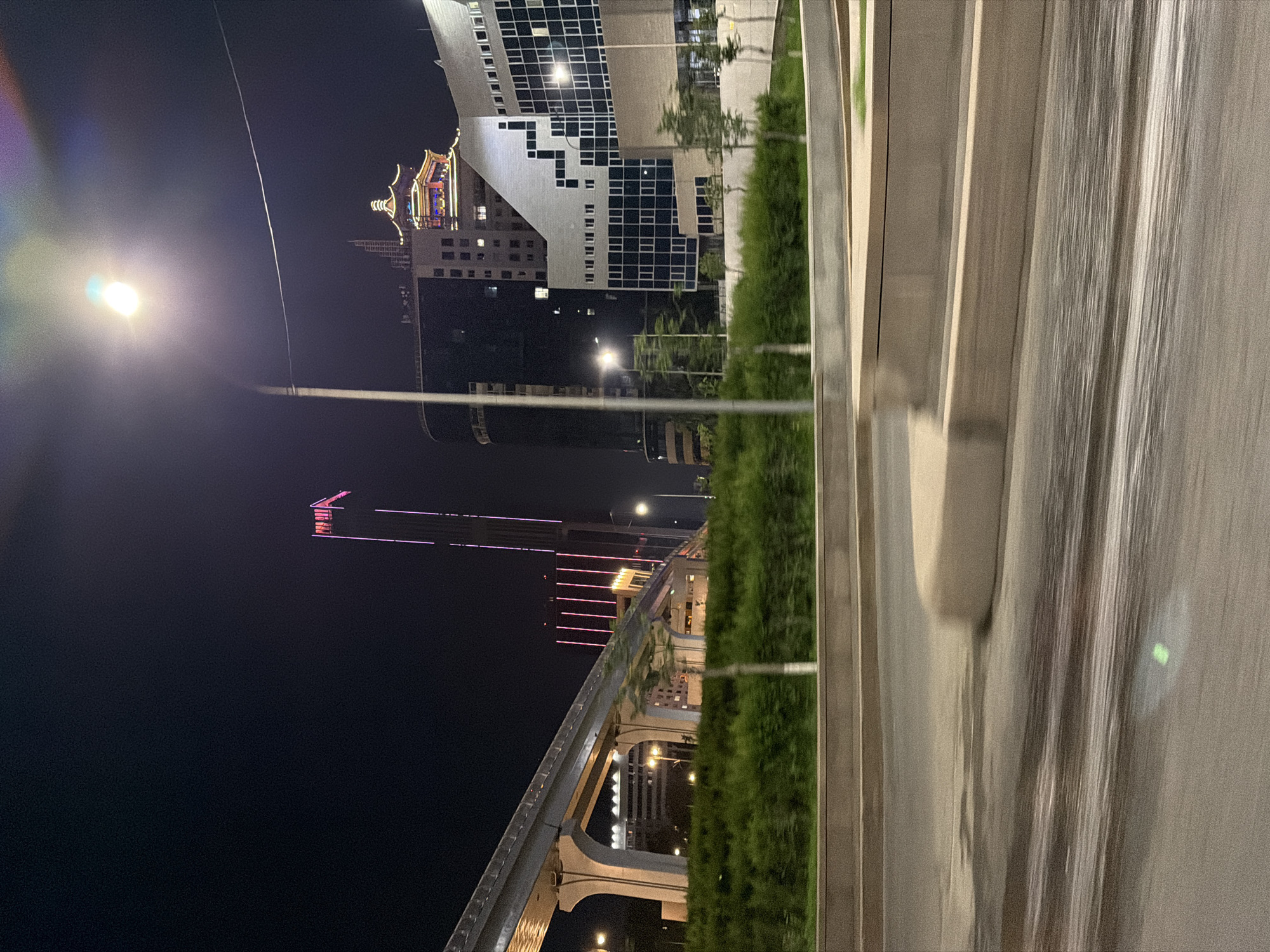The image captures a nighttime city scene in Astana, Kazakhstan. In the foreground, a blurred road surface suggests movement, possibly from a vehicle. A concrete barrier with a curved top is visible in the mid-ground. Beyond this, a dense green hedge line runs horizontally across the frame. Above the hedge, an elevated structure, likely a monorail or sky bridge, is illuminated, supported by large concrete pillars.
In the background, several buildings are visible. A tall, slender skyscraper with vertical pink lighting is prominent on the right side. To its left, a building with a traditional East Asian-style roof, illuminated with colorful lights, sits atop a darker, modern structure. Another modern building with a distinctive white geometric facade and a grid of dark windows is also present. A street lamp with a bright light is positioned in the upper right quadrant, casting a glow and creating lens flare. The sky is dark, indicating nighttime. No people are visible in the image.