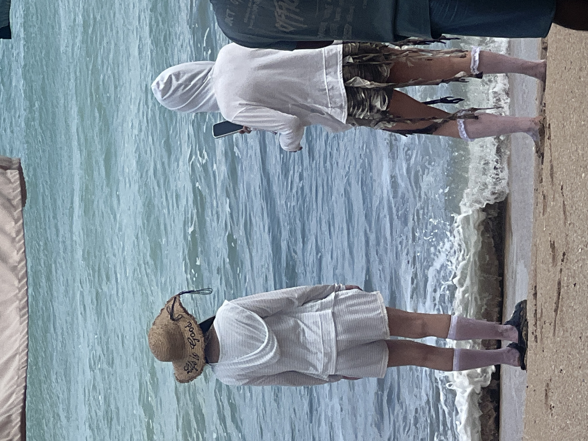 Two individuals are standing on a sandy beach facing the ocean. The person on the left is wearing a straw hat with the words "Life is Good" embroidered on it, a white hooded long-sleeved shirt, white shorts, white socks, and dark sandals. The person on the right is wearing a white hooded long-sleeved shirt, distressed patterned shorts, and light pink socks. The person on the right is holding a mobile phone. The ocean in the background has moderate waves. The image appears to have been taken in Kuta, Indonesia.